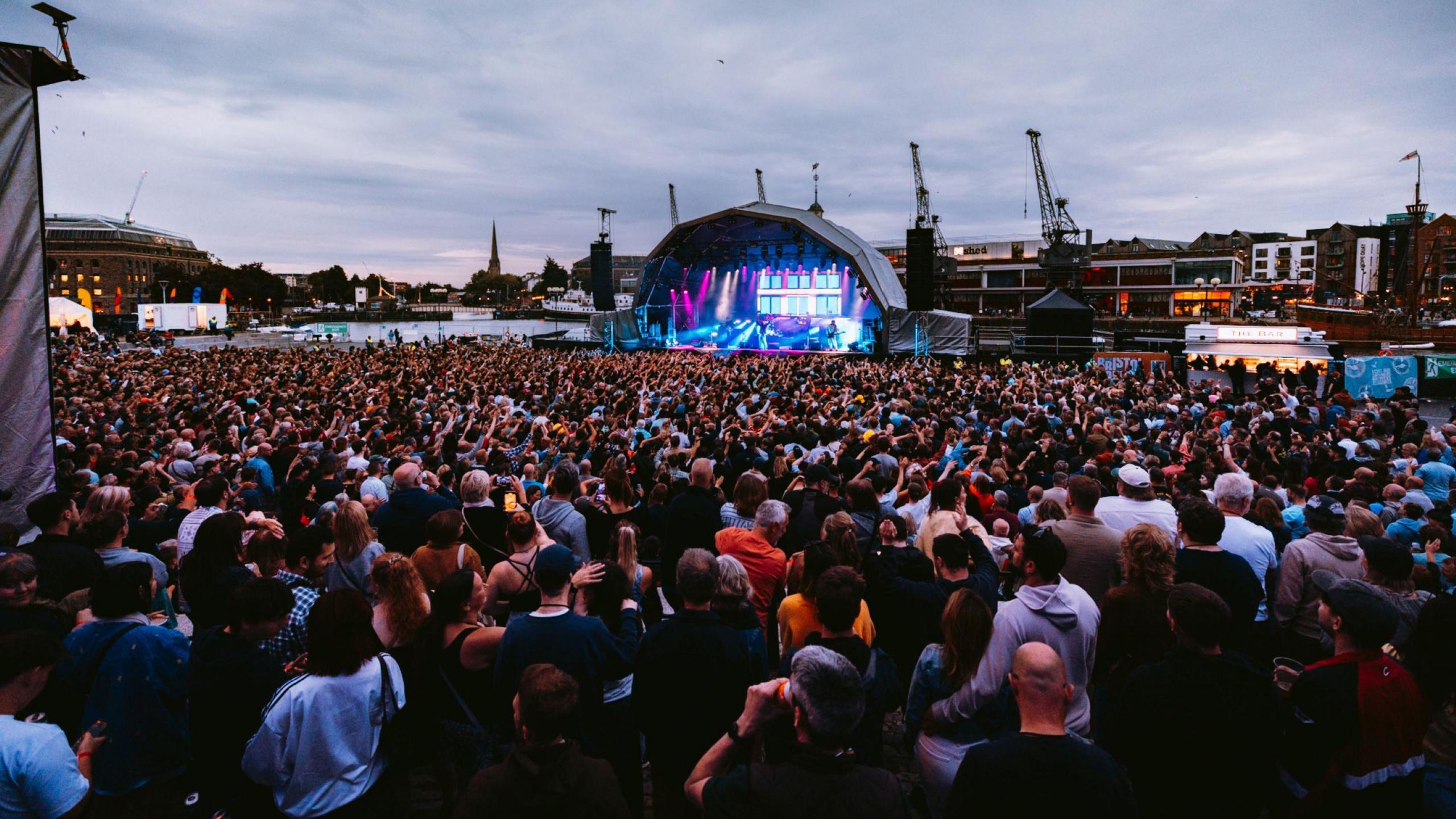A large crowd is seen at night from the back of the Lloyds Amphitheatre during the Bristol Sounds Kaiser Chiefs concert. It is nearly dark and the stage is brightly lit.