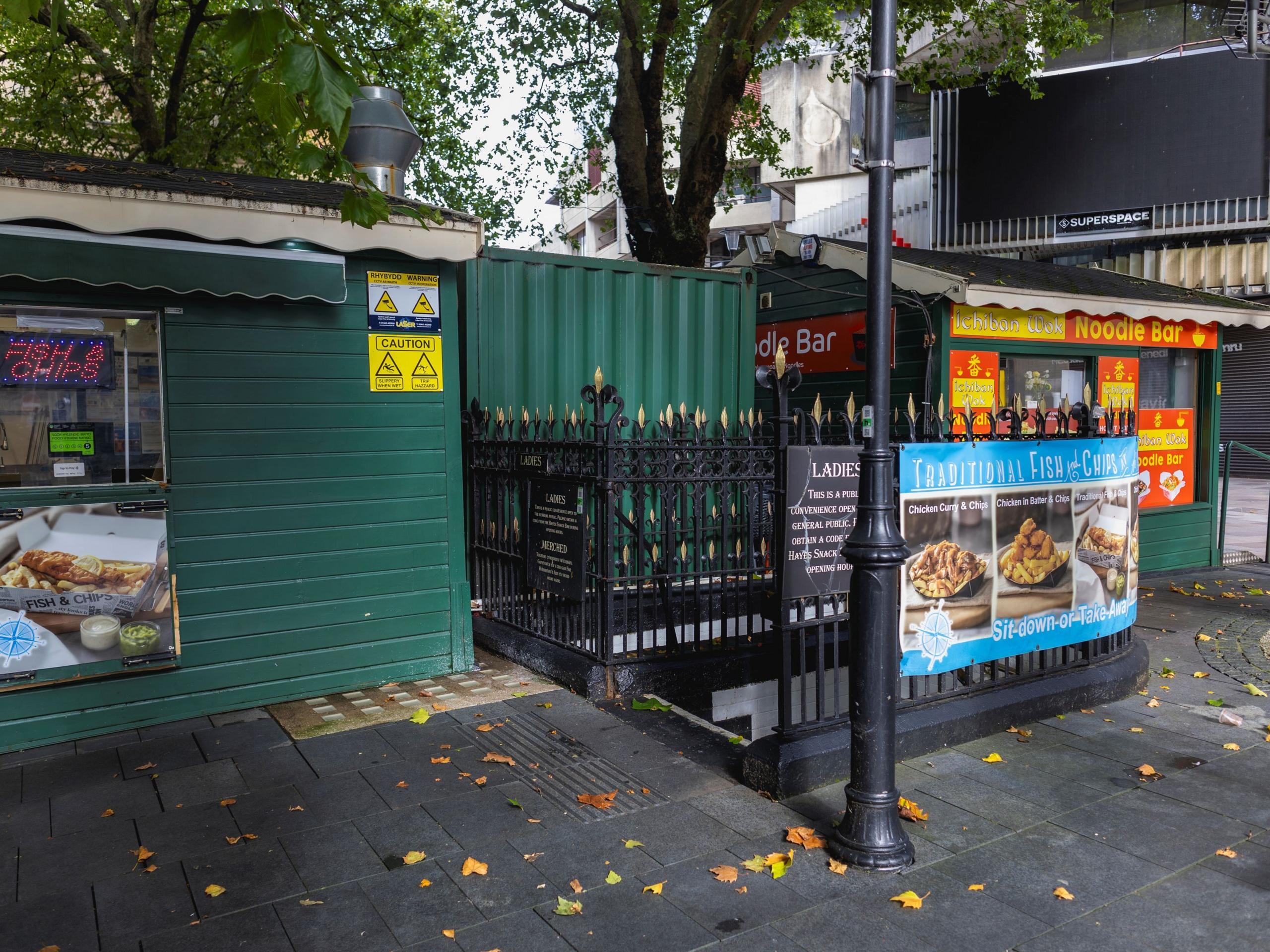 The exterior of a subterranean public toilet. Black railings surround concrete steps on a street next to a green hut selling fish and chips and one selling noodles.