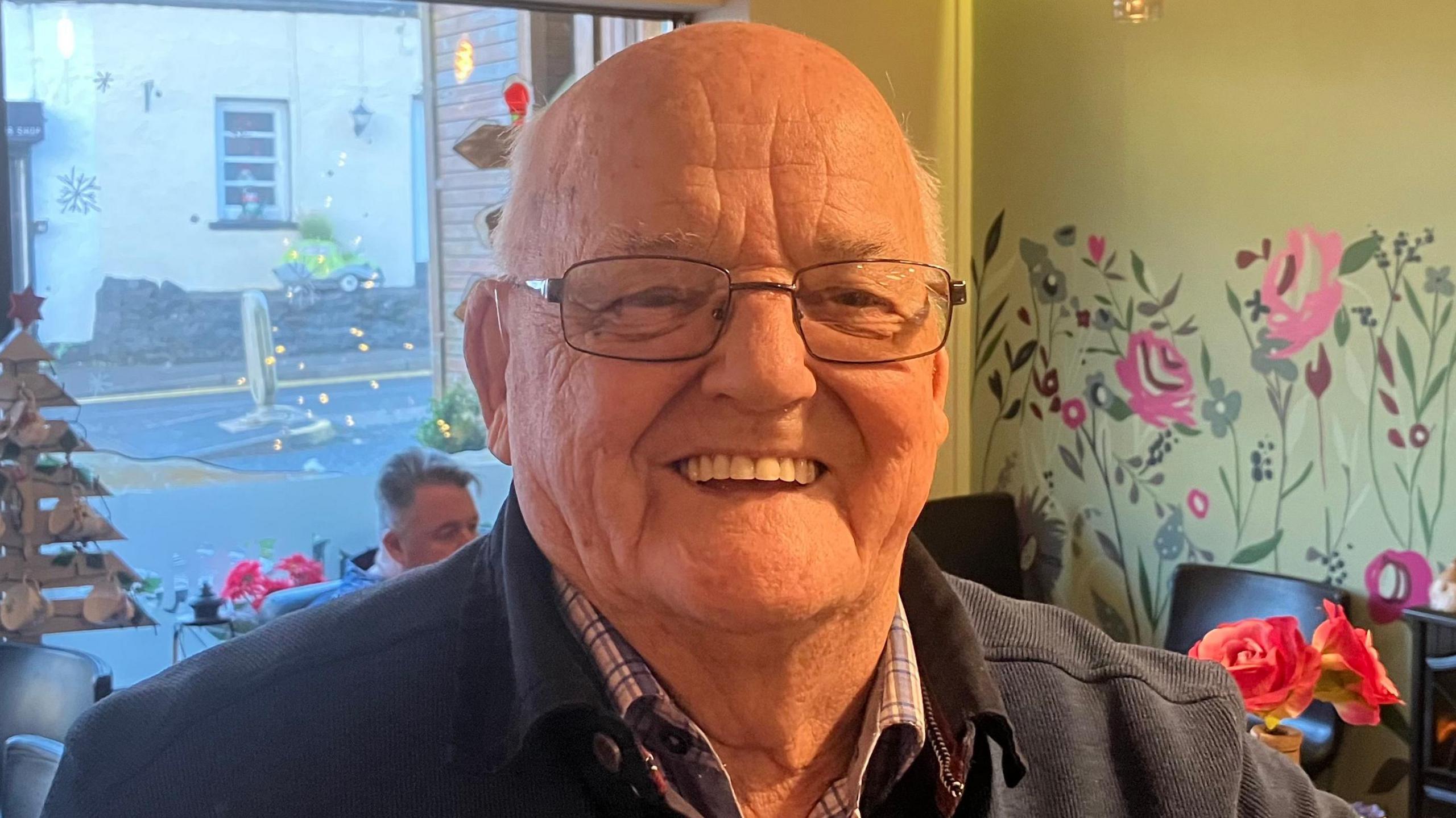 A man sitting on a high stool in a cafe is smiling into the camera. He's wearing a dark grey jumper over a checked shirt.  Behind him in the cafe are tables and chairs and the front window looking out on to a quiet country high street.