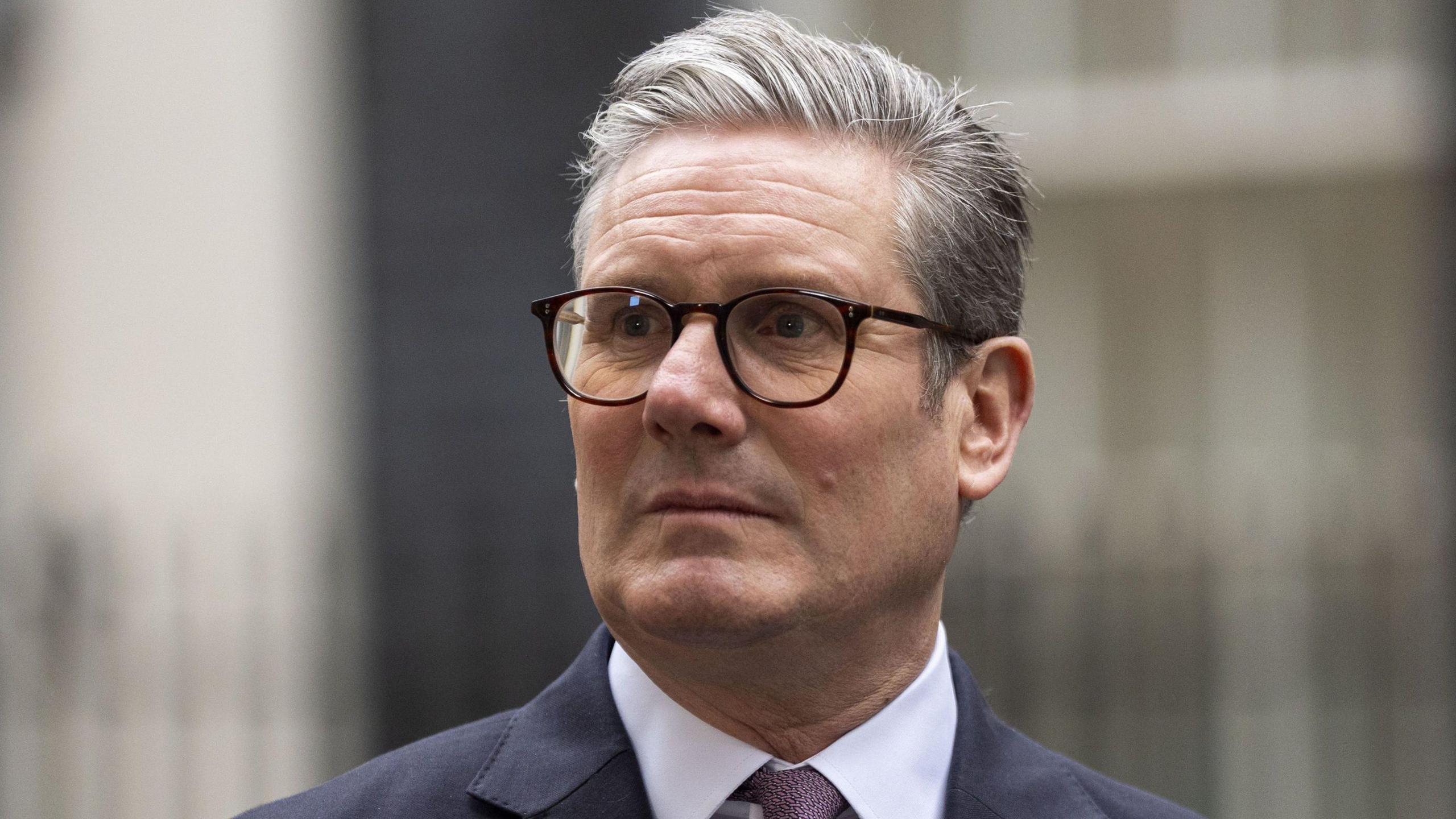 Sir Keir Starmer is looking away from the camera. He has short grey hair and is wearing glasses. He is wearing a white shirt and tie and suit jacket. 