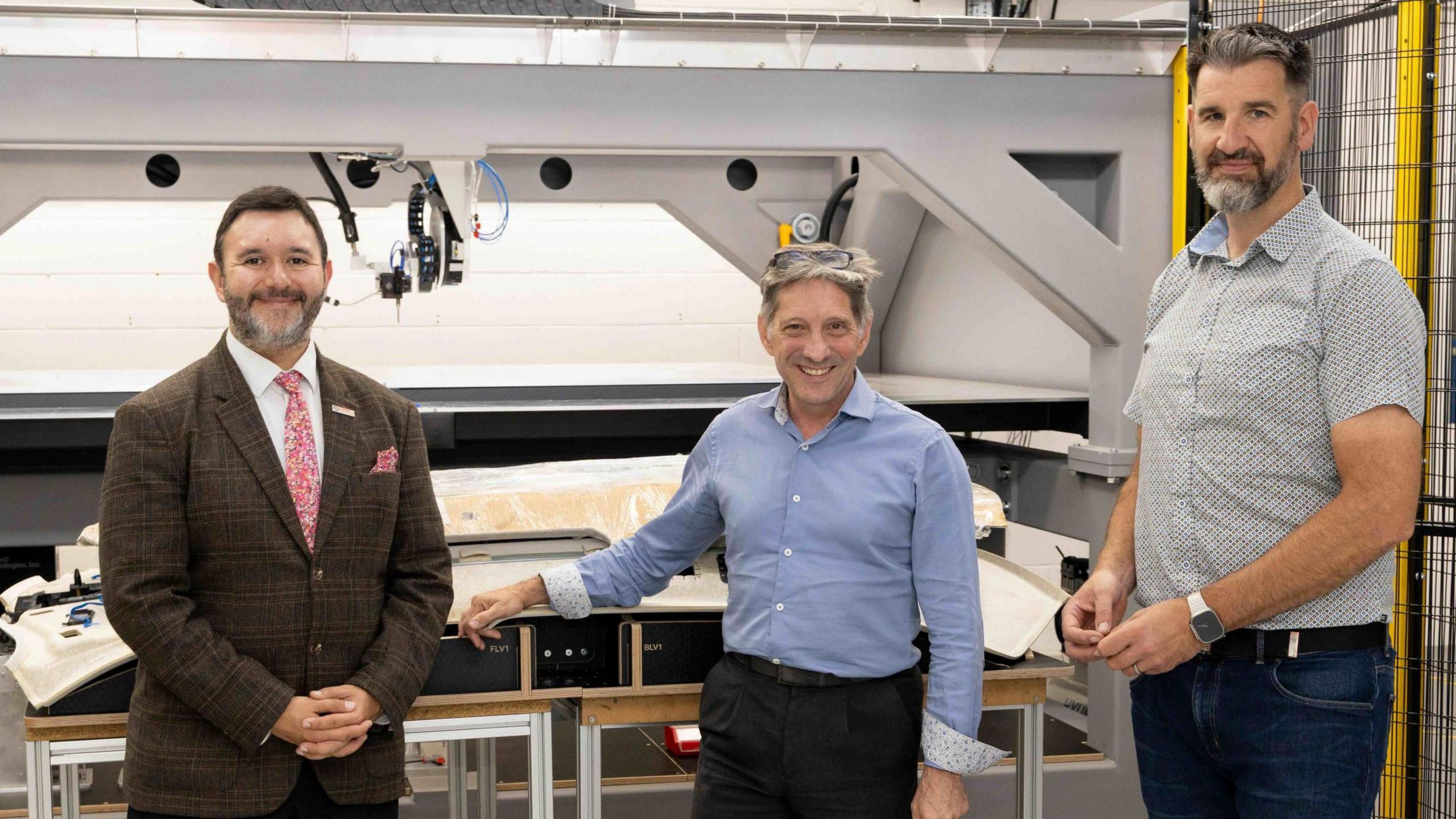 Three men with beards, one of them wearing a suit, stand by some heavy machinery in a warehouse