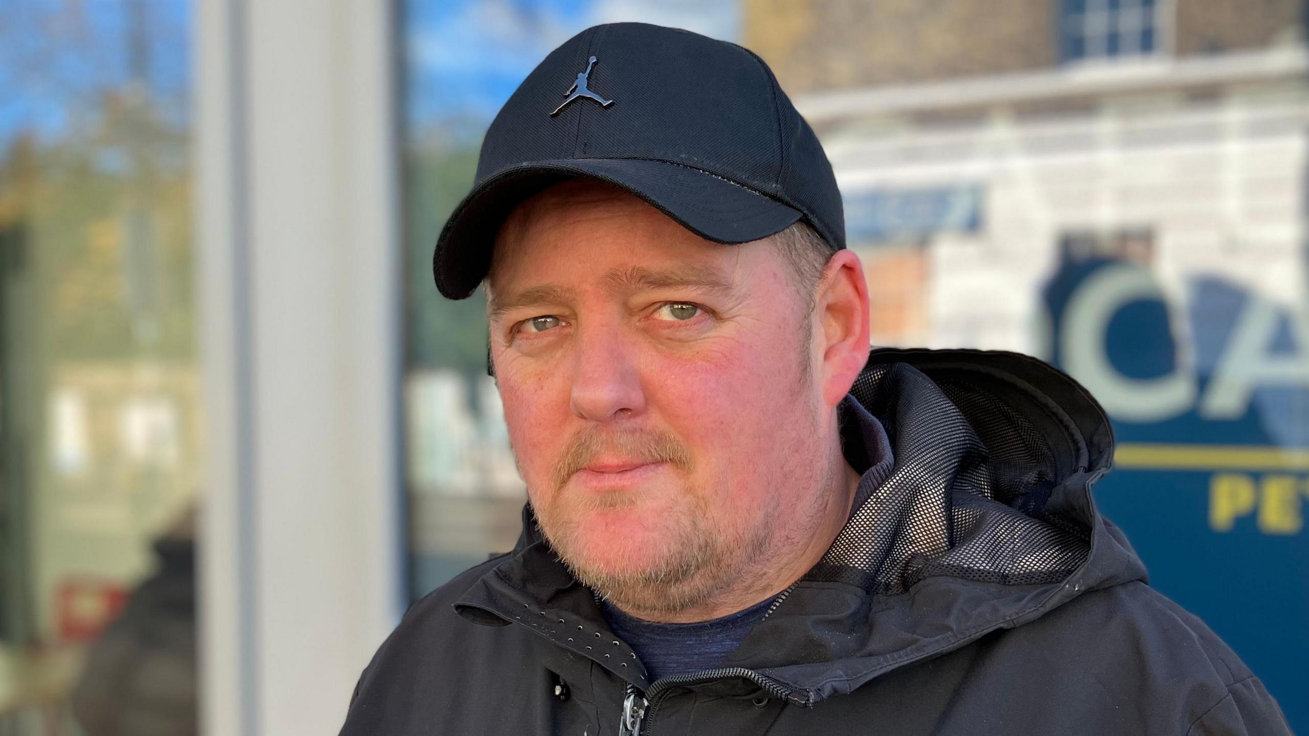 Dal Hicks is in the middle of the image looking at the camera. He is wearing a black baseball cap and a black coat. 