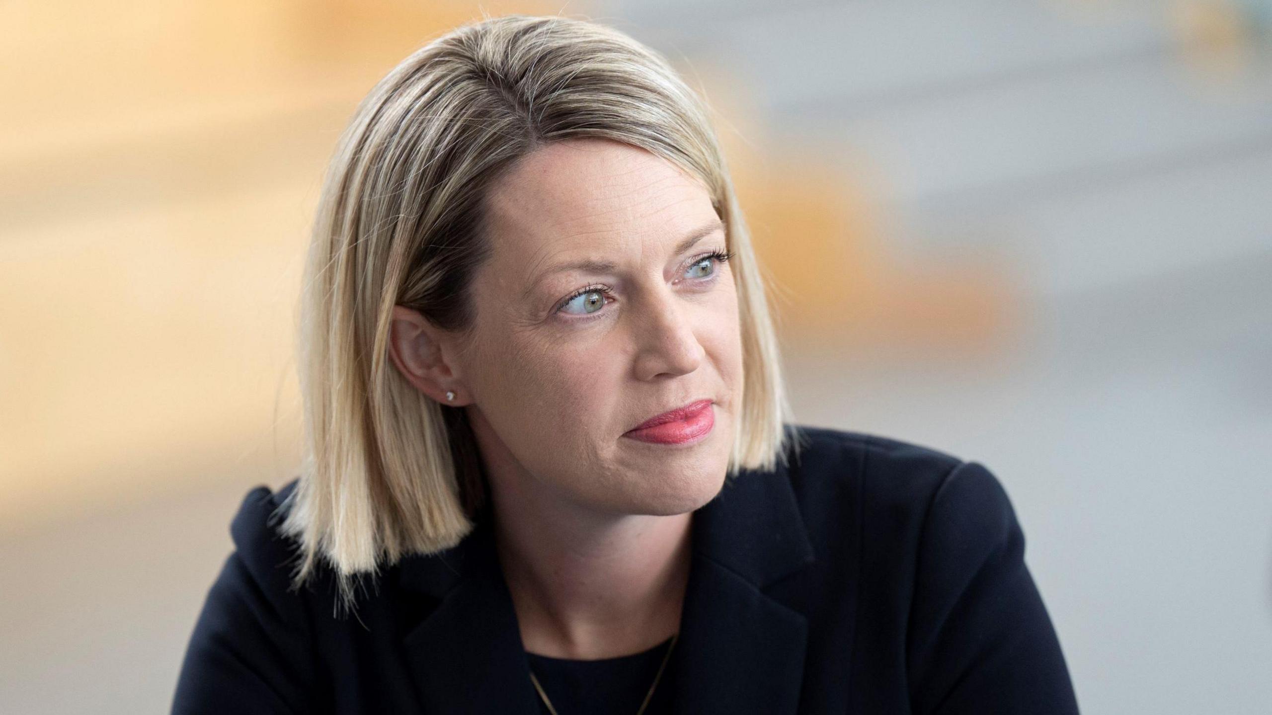 Jenny Gilruth - a woman with blond hair and wearing a dark jacket, looks to her left with a smile on her face