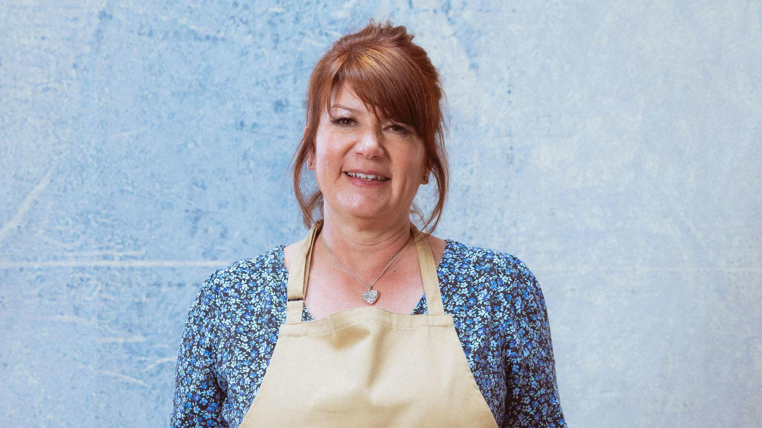 Lesley, a middle-aged woman with brown hair, poses with a blue floral top and a brown apron.