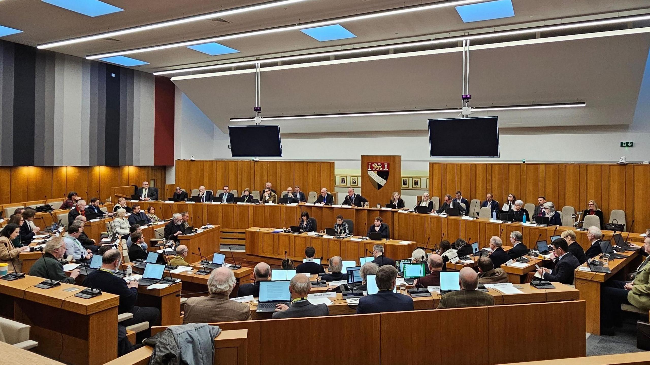 A wide shot of the Norfolk County Council chamber, full of councillors. It is a new modern room, with two large TV monitors in the picture.
