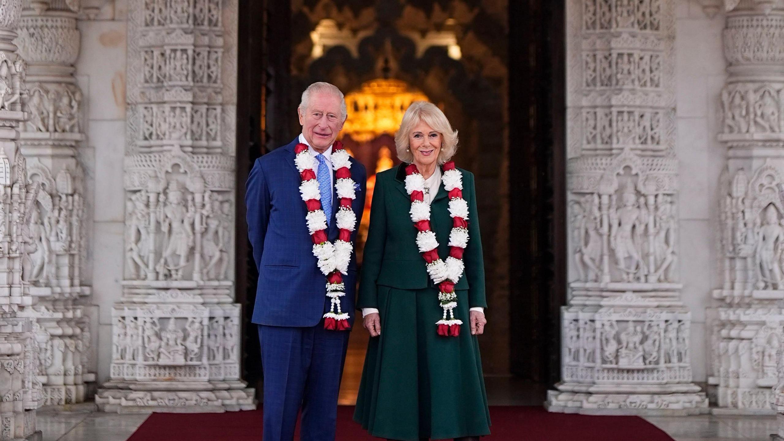 King Charles and Queen Camilla stand in front of a white marble doorway which is decorated with intricate carvings. The couple are both wearing red and white garlands and dark clothes.