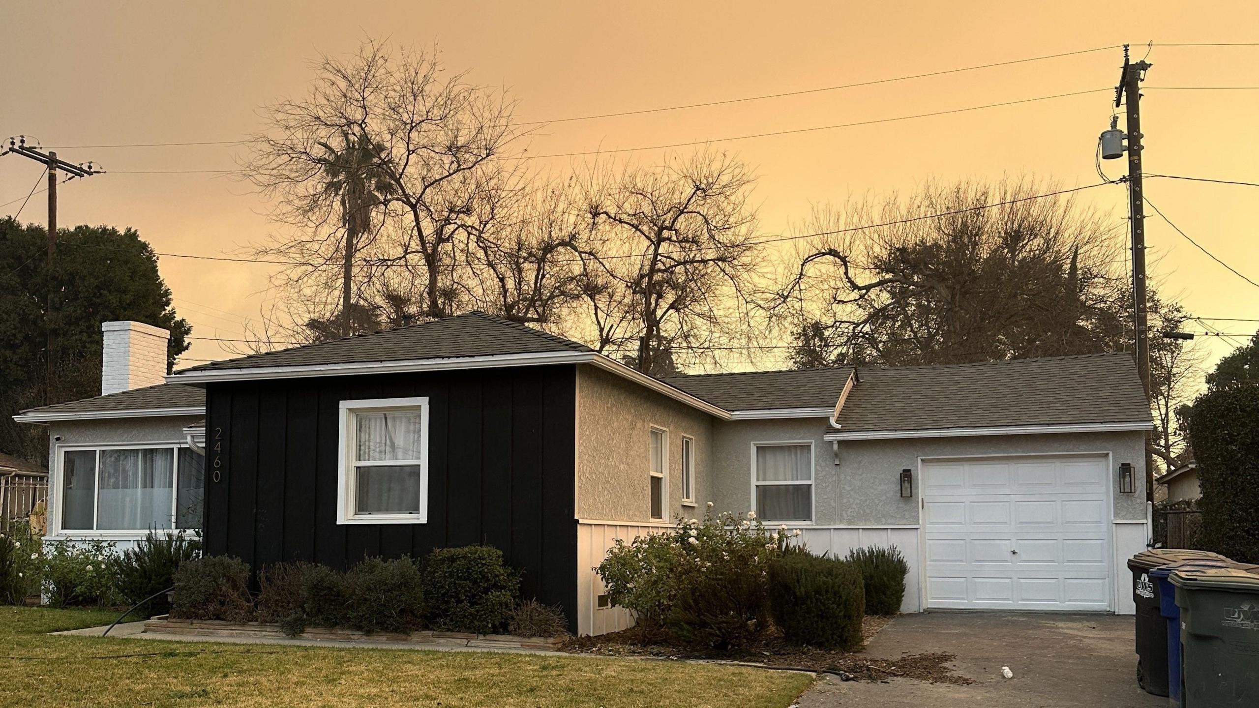 A bungalow surrounded by bushes, trees and a front lawn in front of a yellow/orange sky.