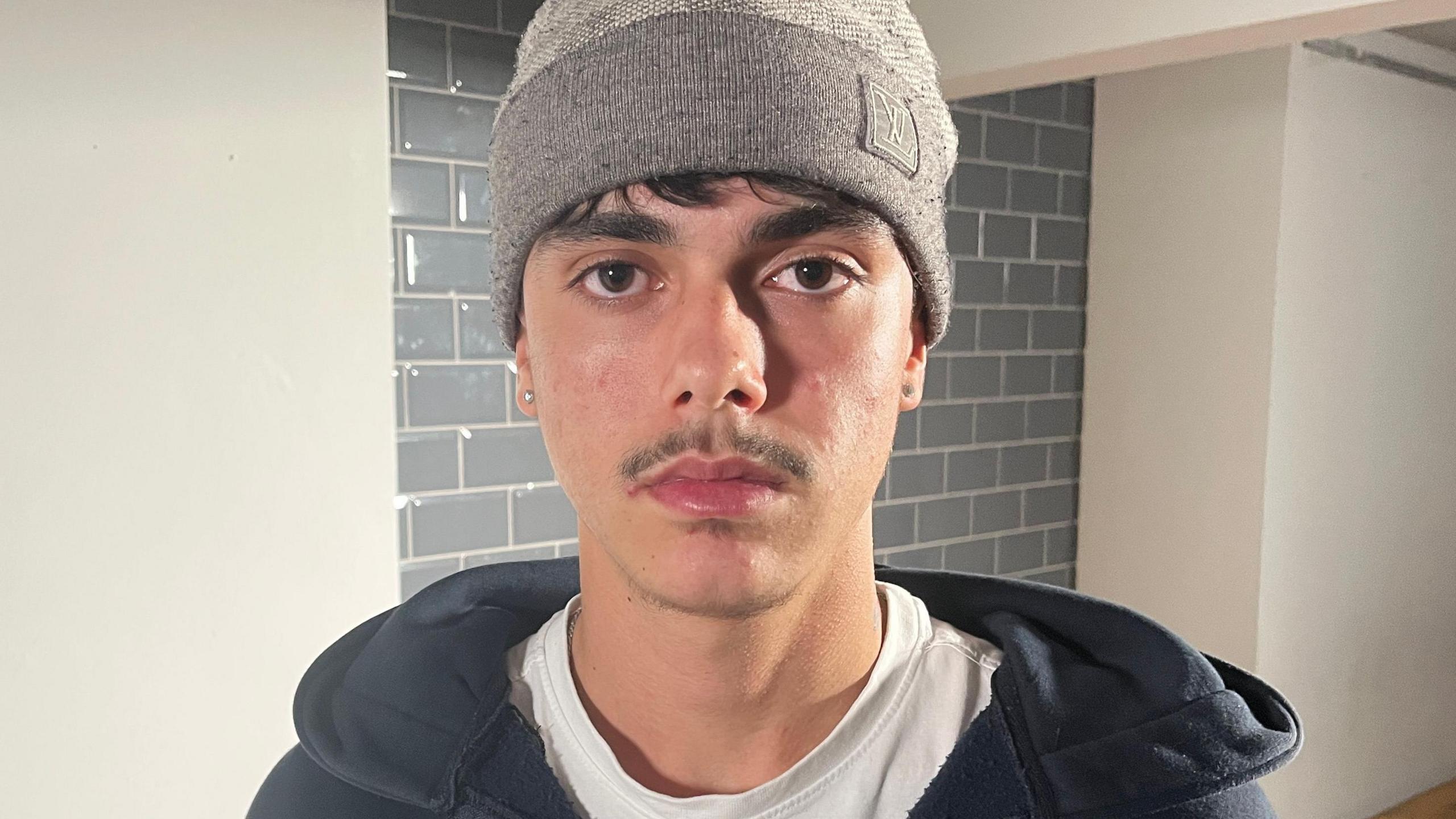 Brooklyn is 18 years old. This is a close up photo of him looking into the camera with a serious expression. He wears a light brown beanie hat. His short, dark hair can be seem sticking out of the brim at his forehead. He has a pencil style moustache and stud earrings in both ears. He wears a white t-shirt with a dark grey hoodie.