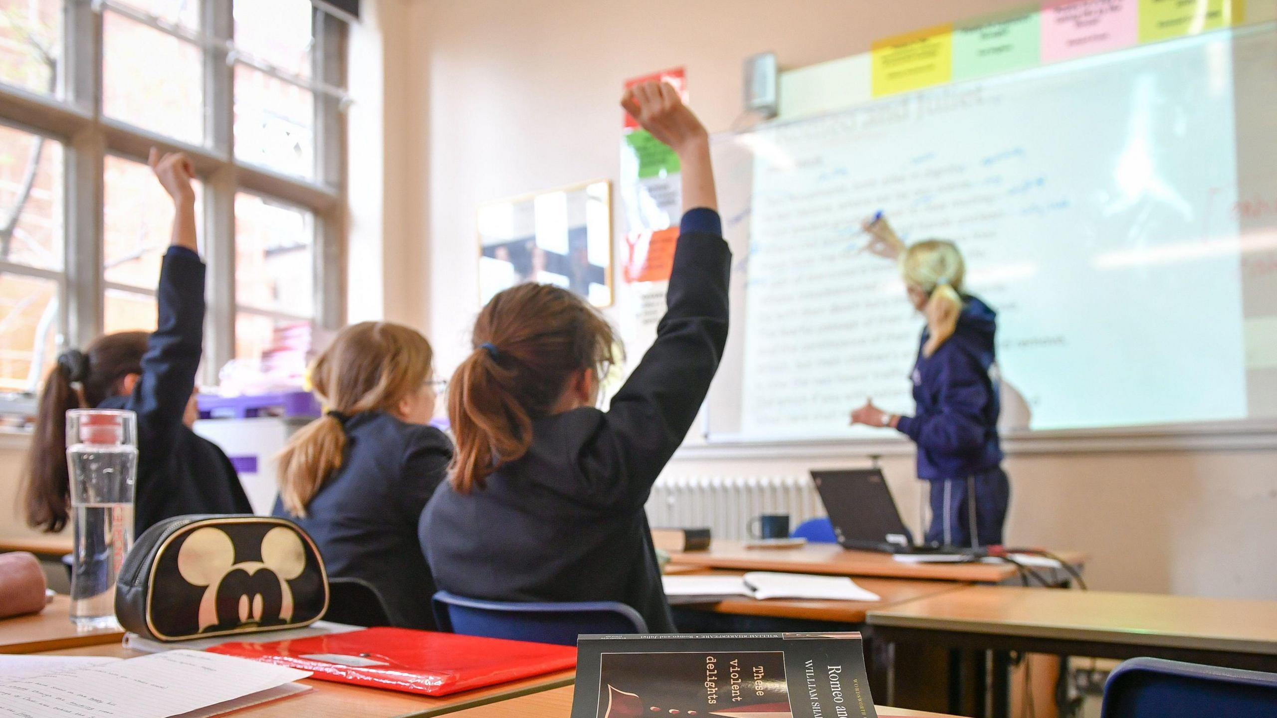 Three pupils sit at a classroom desk while a teacher writes on a whiteboard in front of them. Two of the pupils have their hands raised.