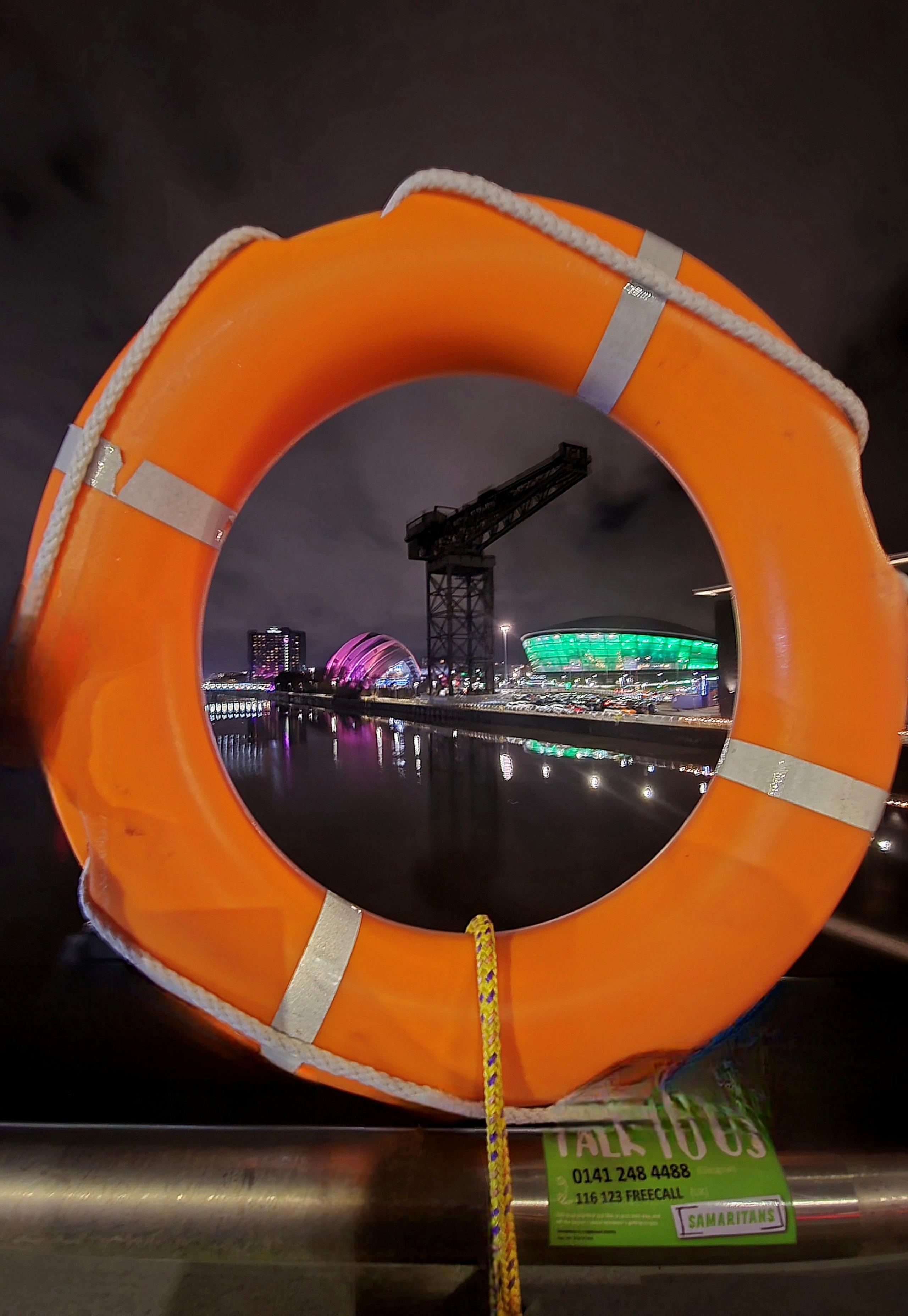 A circular orange life ring on the riverside, with a large crane and large arena visible through the space in the centre of the aid