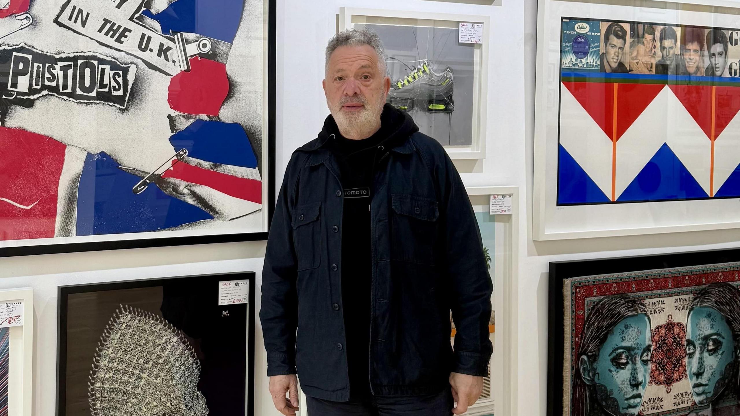 A man with white hair and beard in a navy coat over a black hoodie. There is artwork on the wall behind him.