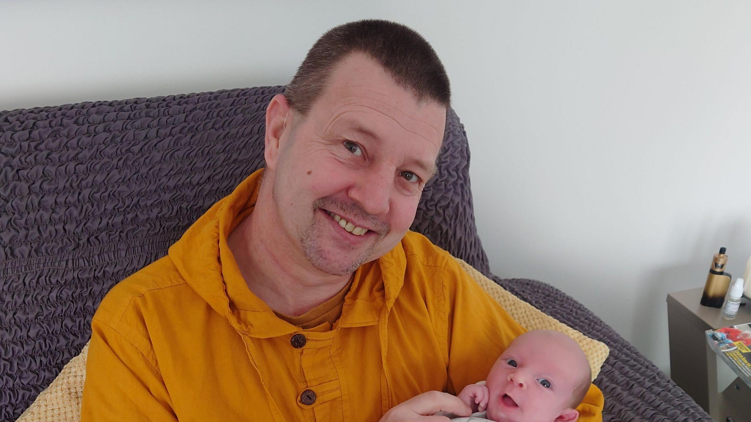 Nathan Clements is smiling at the camera, wearing an orange hooded top and with a baby in his arms. 