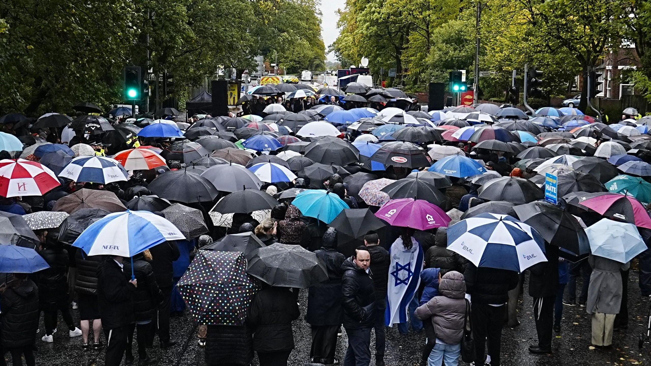 A large crowd of people, many holding umbrellas, stand in front of a stage flanked by stacks of large speakers. 