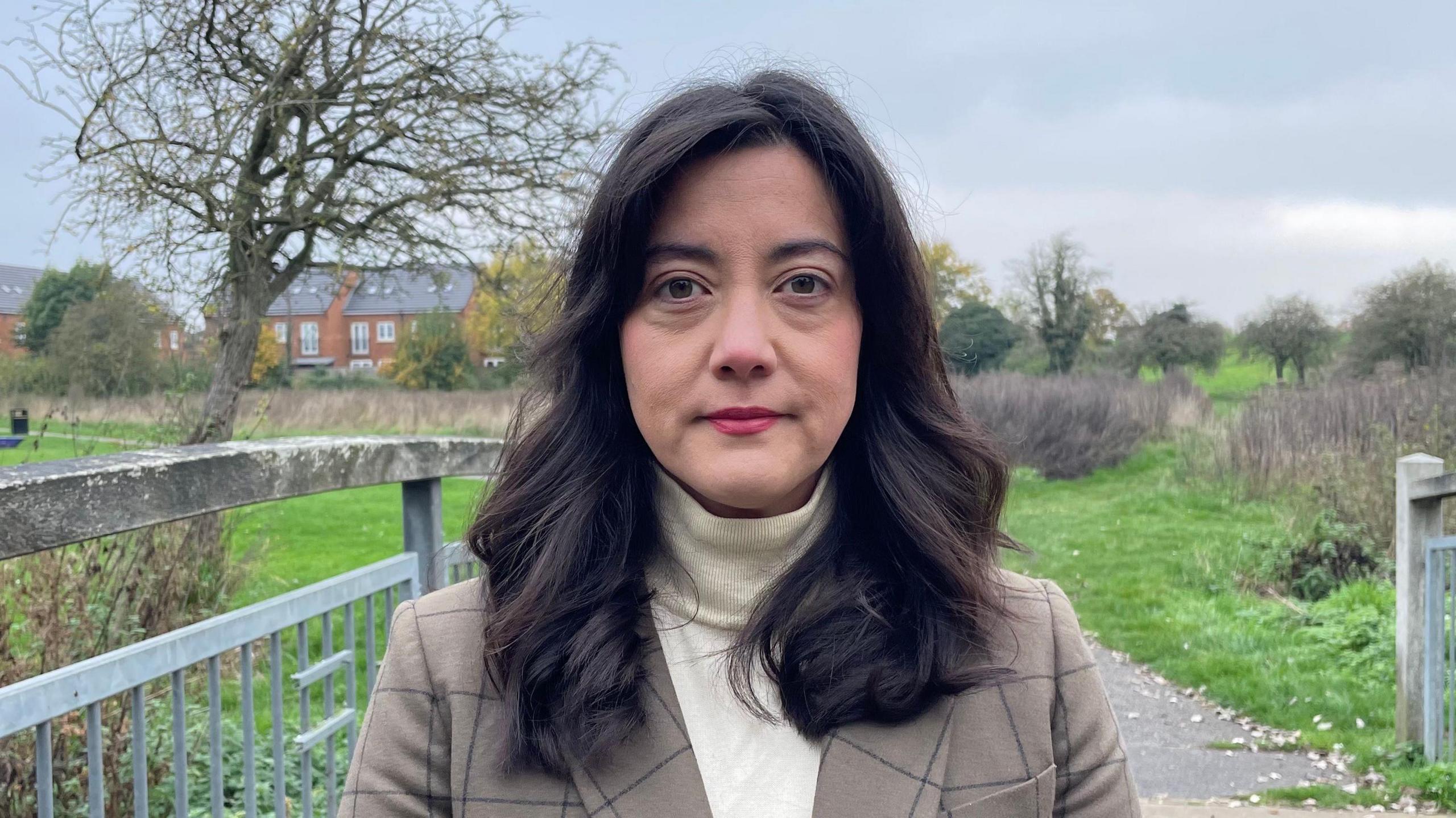 Sarah Owen looking direct to camera. She is standing outside in an open space, with grass, trees and some housing in the background. To her left are fence-type railings. She is wearing a light brown jacket and cream high-necked top. She has shoulder length dark drown hair and brown eyes. 