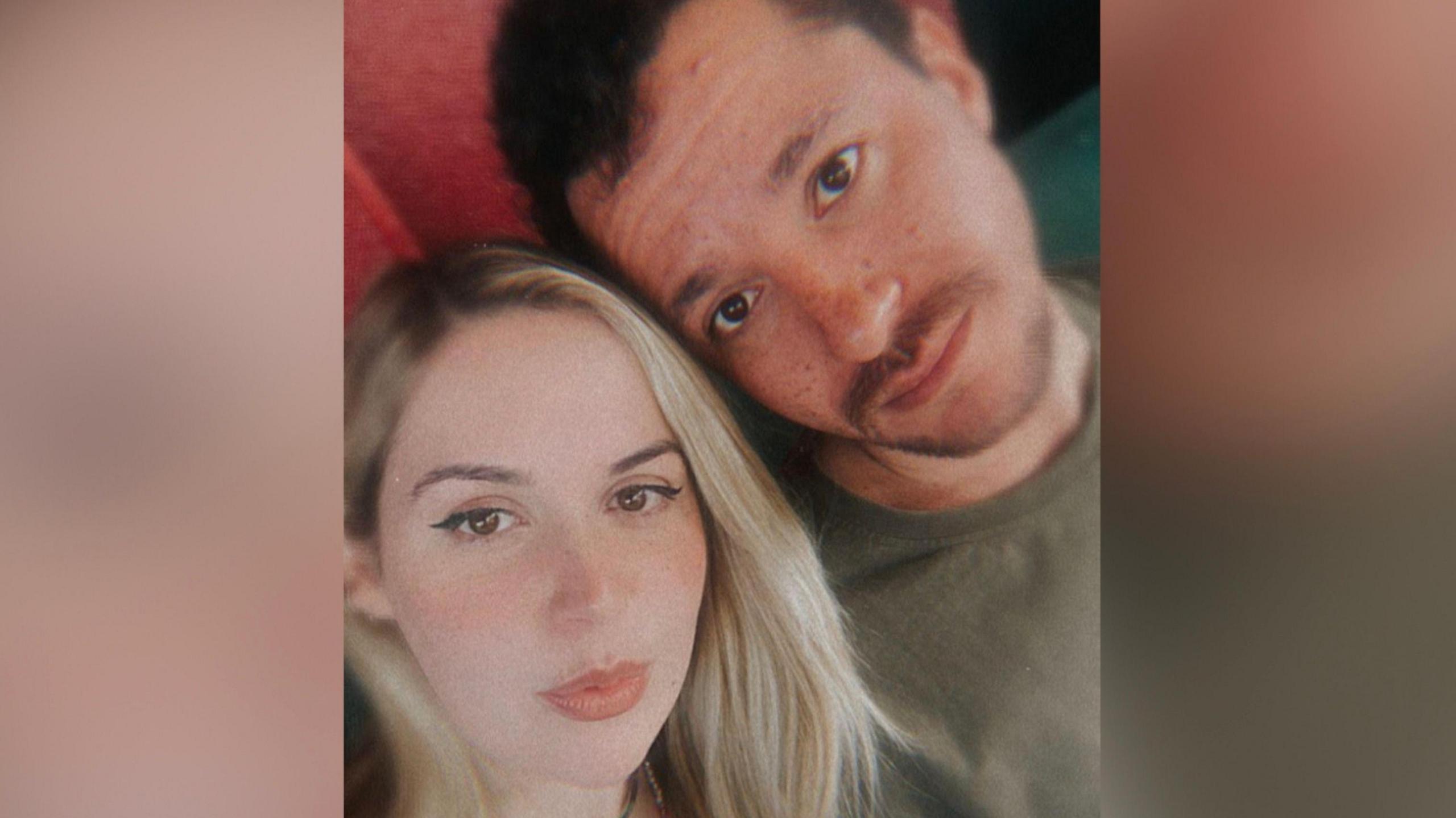 A woman with long blonde hair is looking at the camera.  She is next to a man with brown hair and a moustache. The sides of the image are blurred.