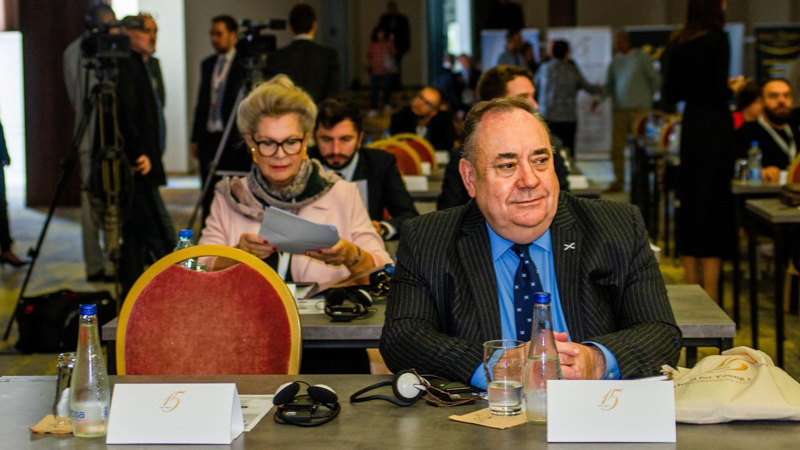 Alex Salmond sitting a table with a white nameplate in front of him. The red chair with a gold frame next to him is empty. Salmond is wearing a dark suit with a blue shirt and dark blue tie, with a Saltire lapel pin.