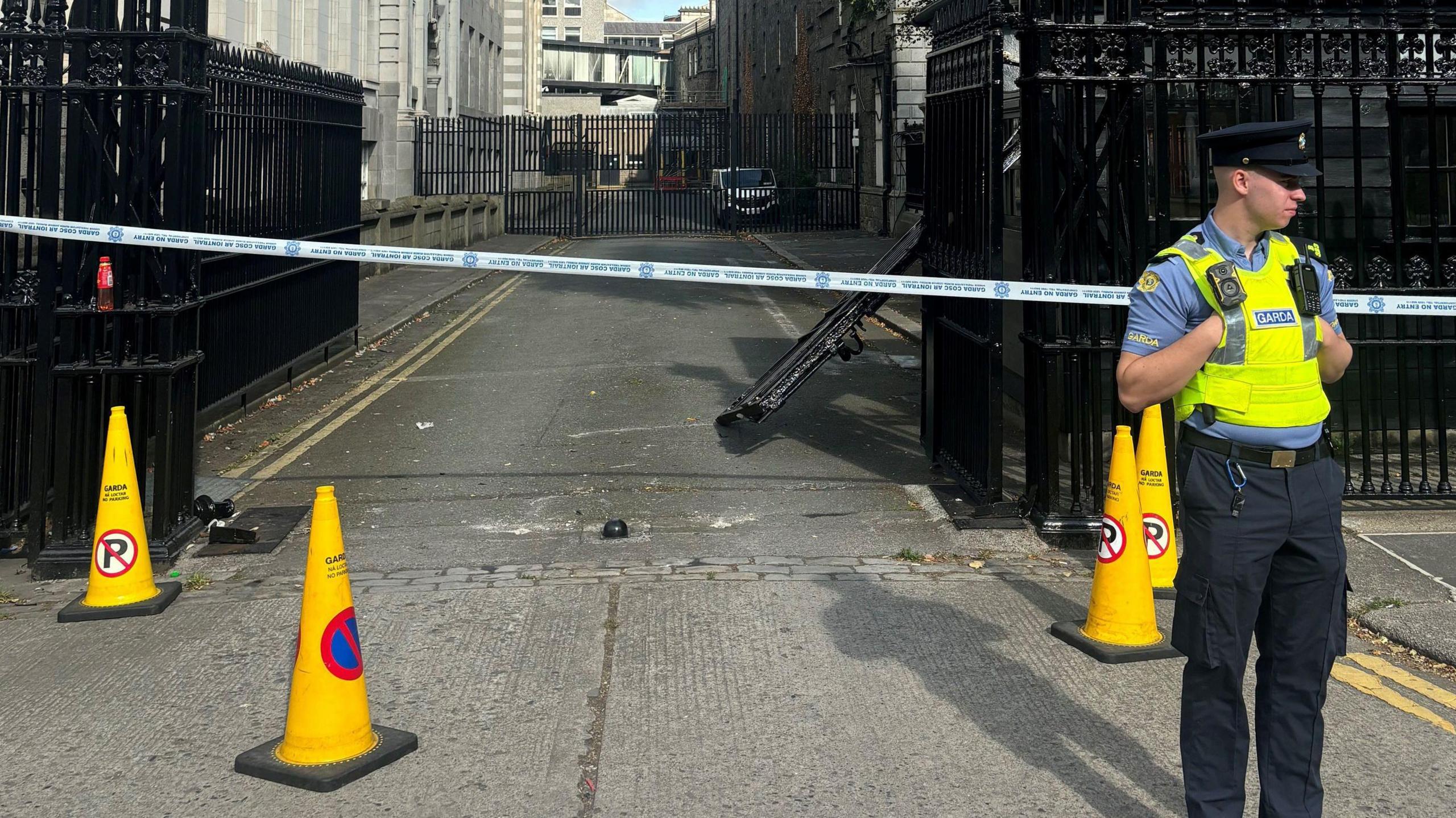 A garda stands with gates behind him. They're been rammed and are broken.