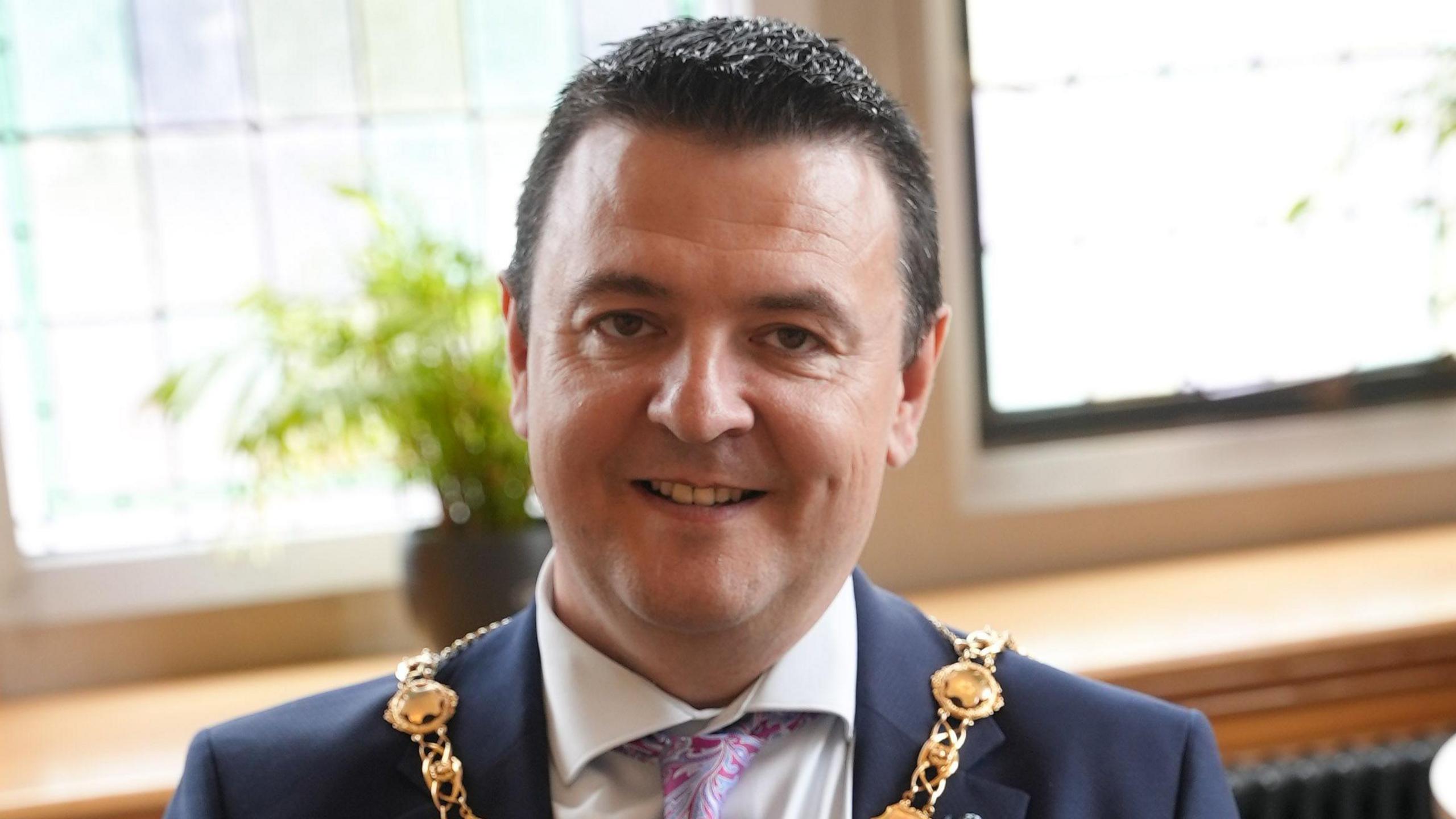 Ruairí McHugh has short black hair and is smiling.
He is pictured in a navy suit and purple and pink tie.
He is wearing his mayoral chain in front of a two window panes.