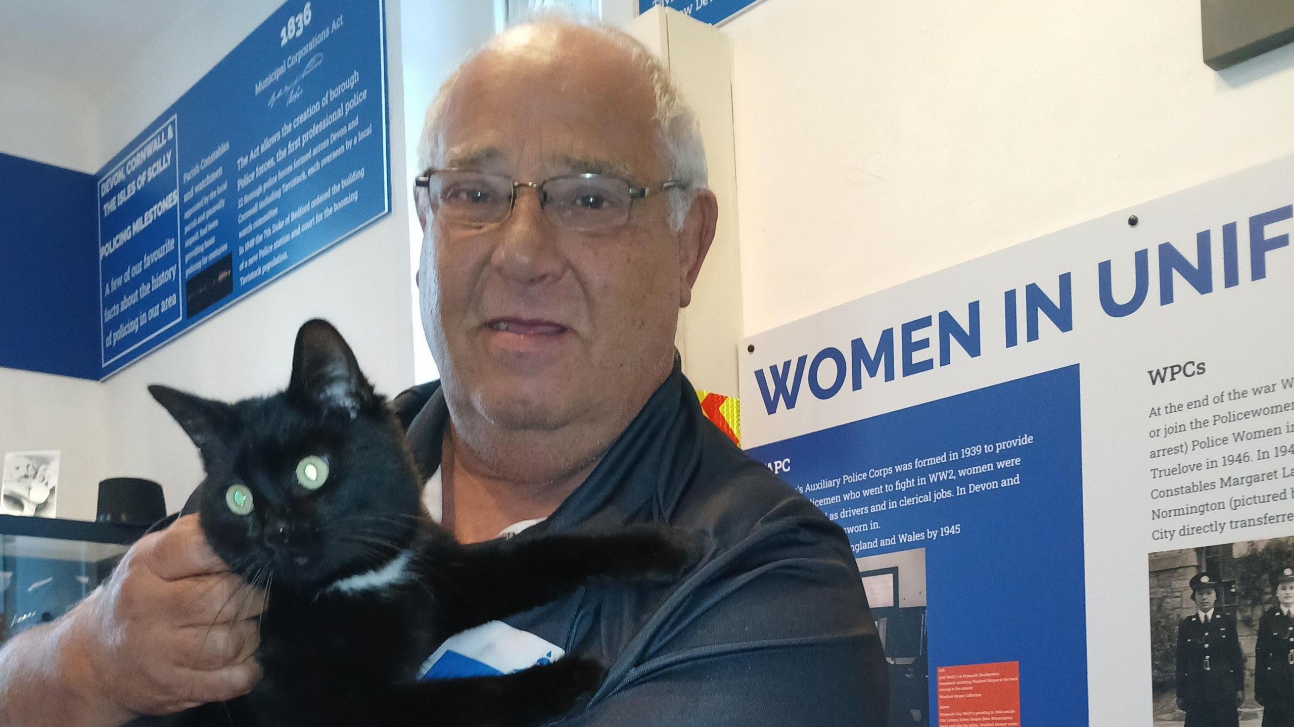 A man holding Polo who was visiting a museum in Tavistock. The man is smiling at the camera and Polo is looking to the left while in his arms. There are signs behind them. 