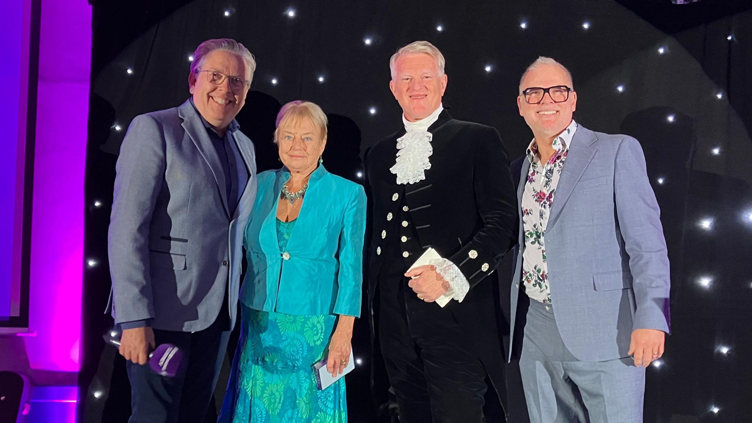 James Cannon, Terri Collins, High Sheriff Peter Cluff and Danny Pike stand on a stage and looking at the camera, smiling. Behind them there is purple lighting and a black fabric backdrop with white lights in it.