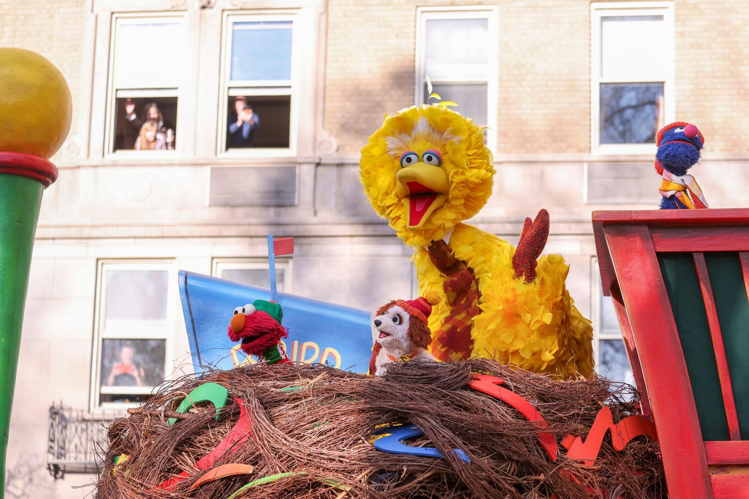 Big bird sits on a nest parade float clapping its hands. People watch out the window behind it.