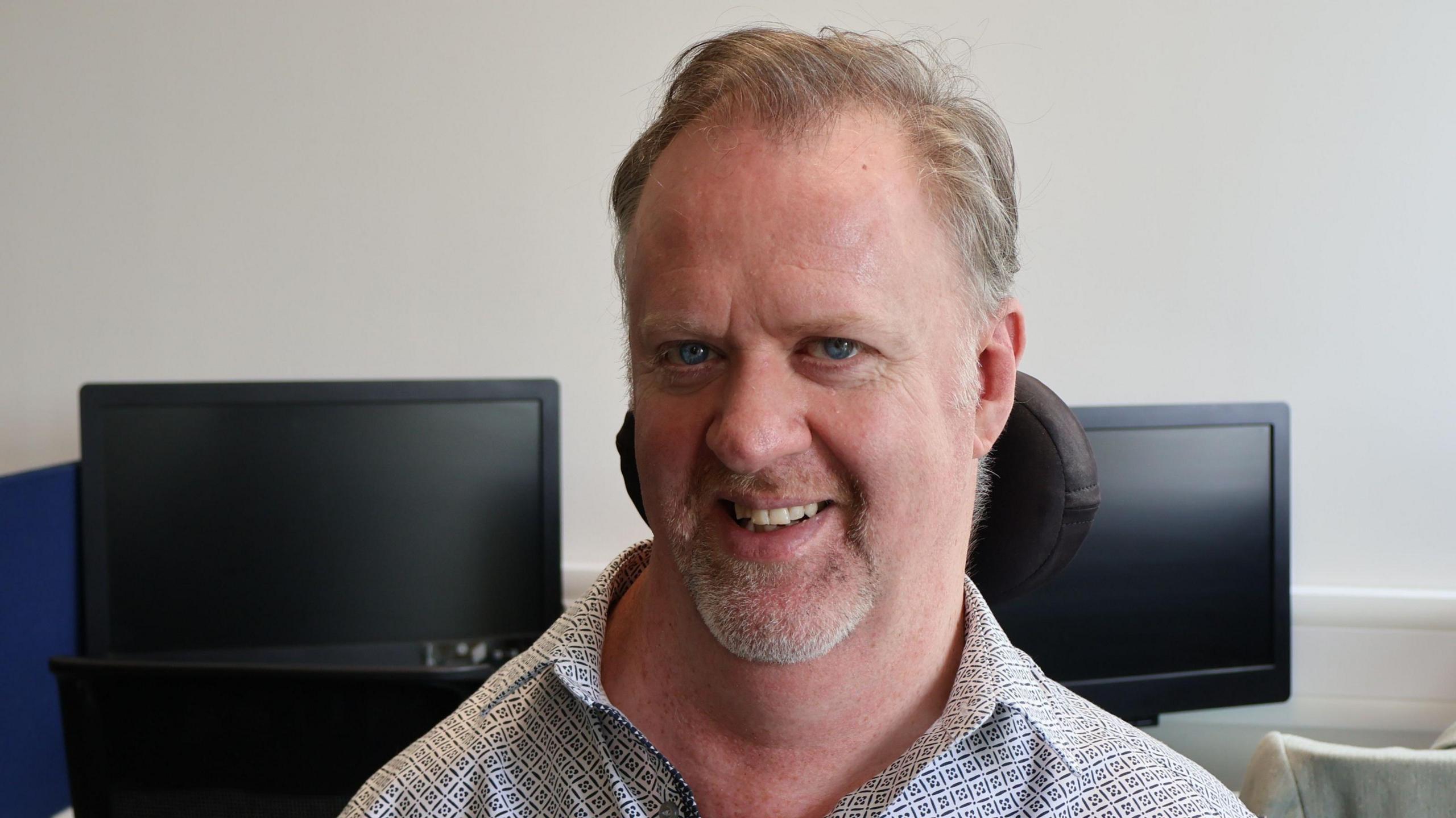 Owen Collumb sitting in a chair wearing white patterned shirt, with two desktop screens in background.