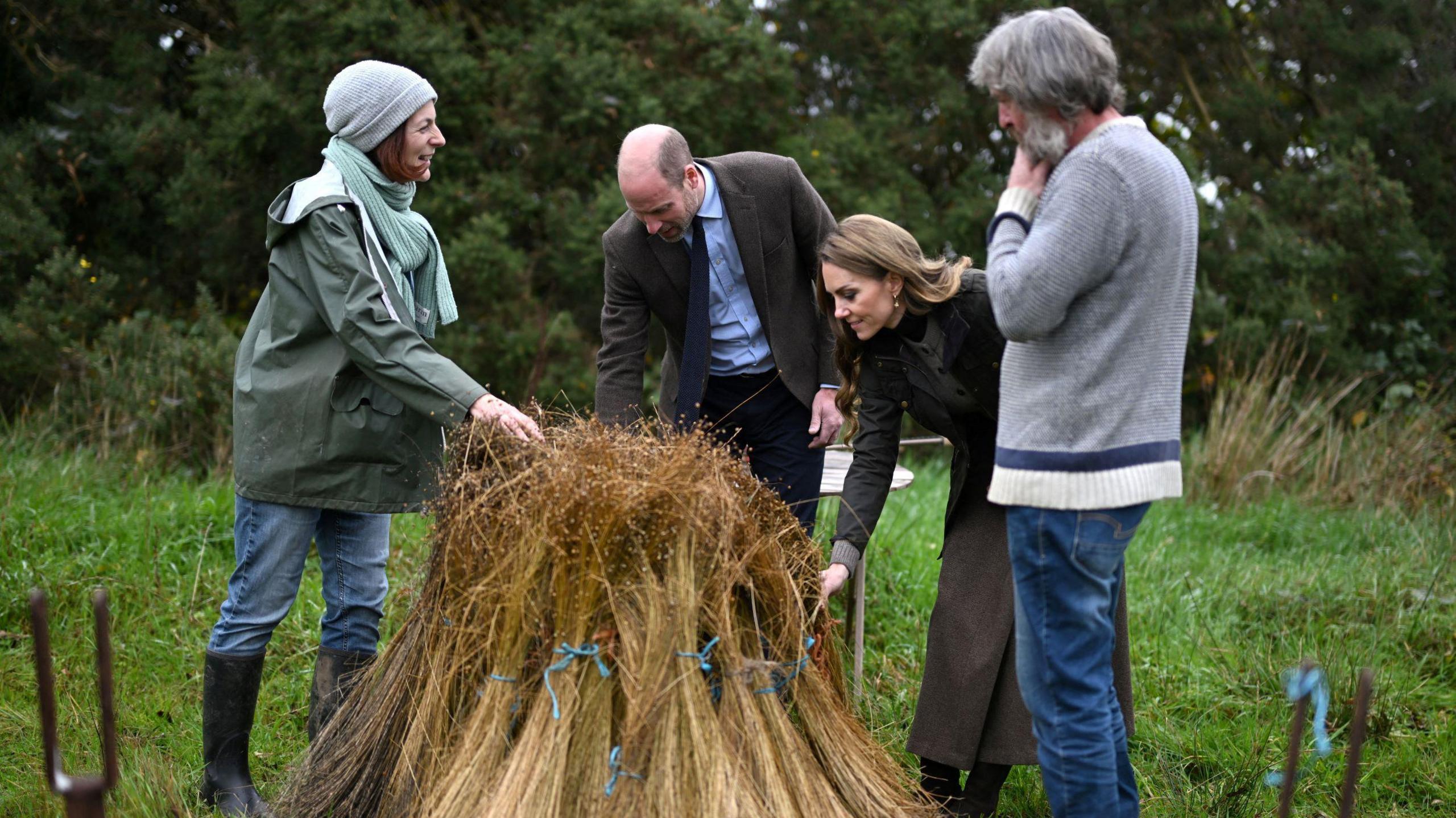 Four people gather round a stack of yellow flax in a green field. The two people in the middle in are the Prince and Princess of Wales. The prince is wearing a brown blazer, blue shirt and navy tie. The princess with long brown hair wears a brown skirt, dark green jacket and brown waistcoat. On the pair's left is a man with medium length grey hair and a short grey beard. He wears blue denim jeans and a grey sweater. On the right is a woman with short brown hair who wears a grey beanie, green waterproof coat, blue jeans and black wellington boots.