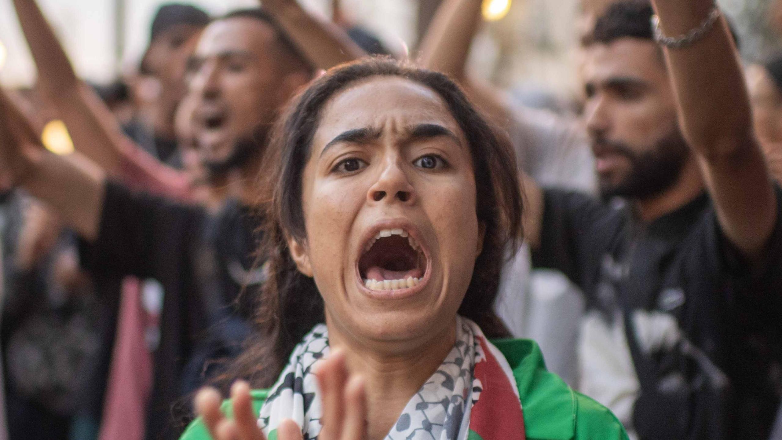 A protester looks at the camera as she chants a slogan. She is wearing a green top and is clapping her hands