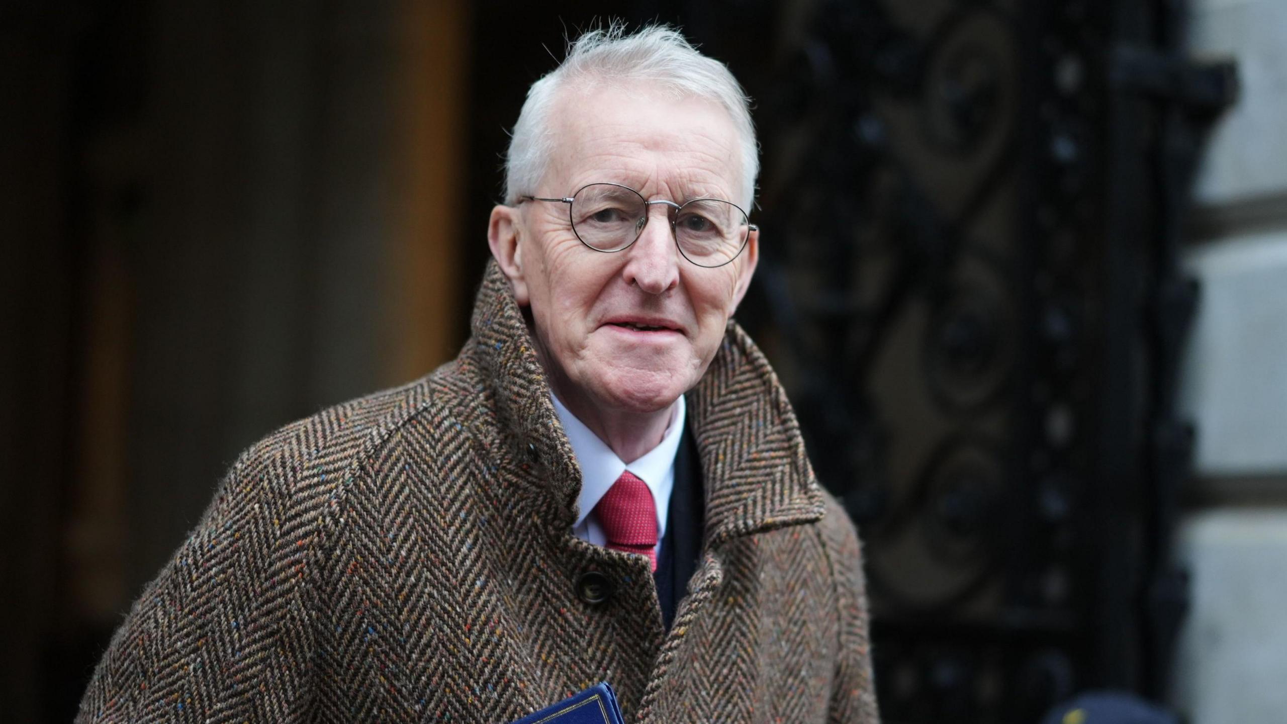 Hilary Benn has light grey hair and metal-framed glasses and is wearing a brown coat over a blue shirt and red tie
