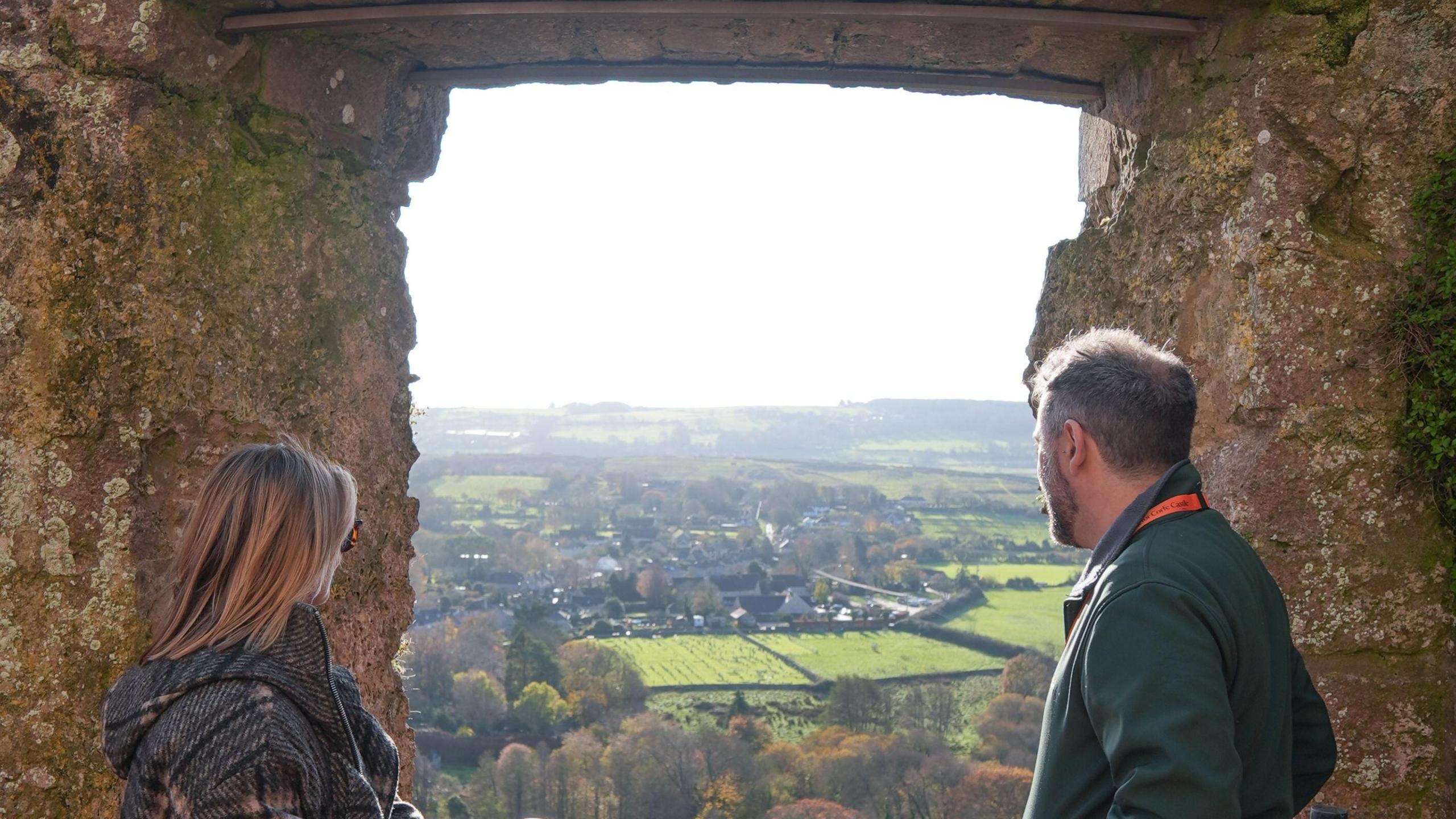 A woman on the left and a man on the right both peering out of a lookout from the old castle walls. Fields, housing and trees can be seen in the distance as they look out on the scene.