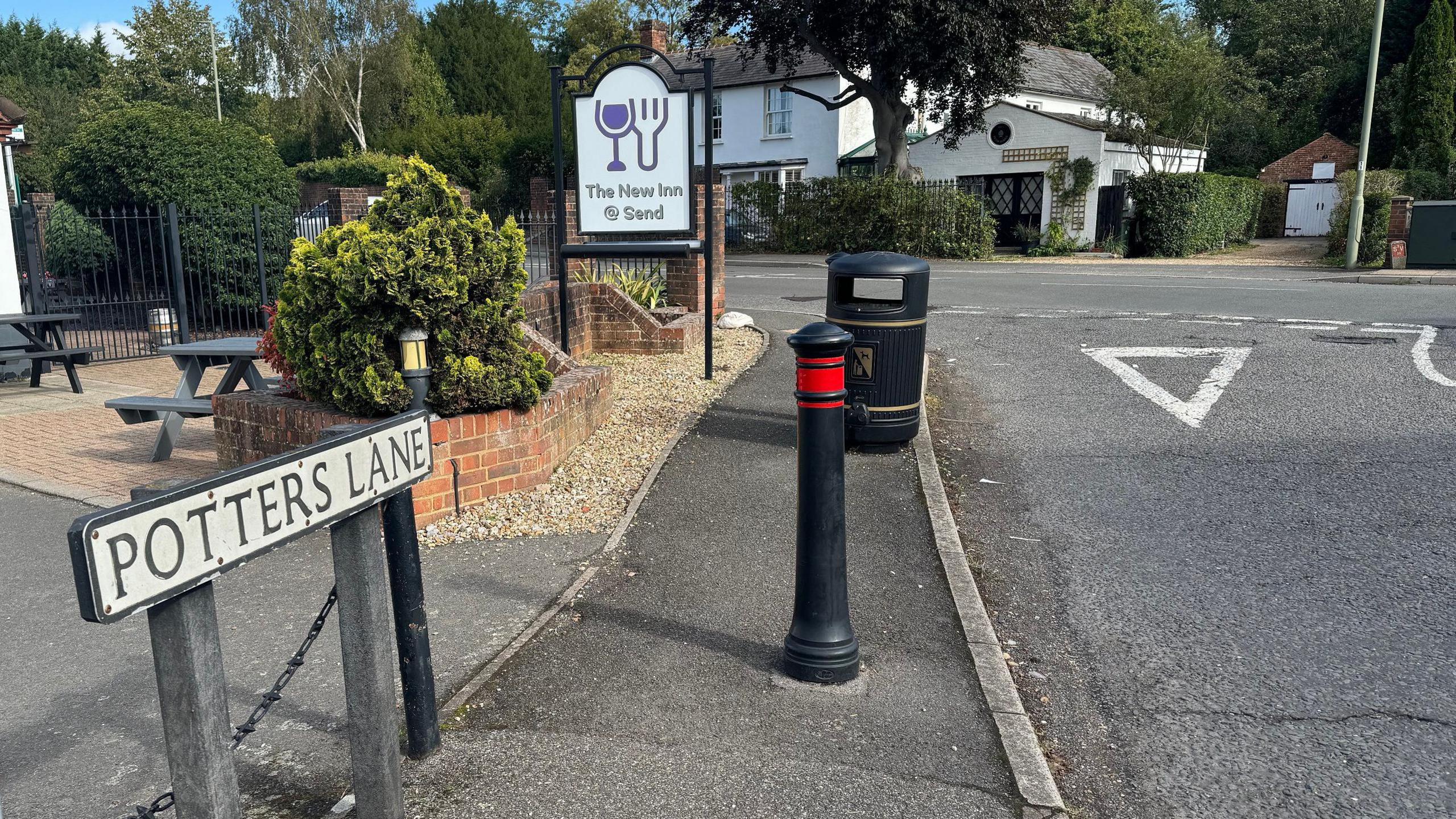 The sign for Potters Lane and then a give way road marking which leads on to the A247 Send Road in Send. In the background there is a sign for the New Inn pub, as well as outside tables for customers. There is also a bollard and a rubbish bin.