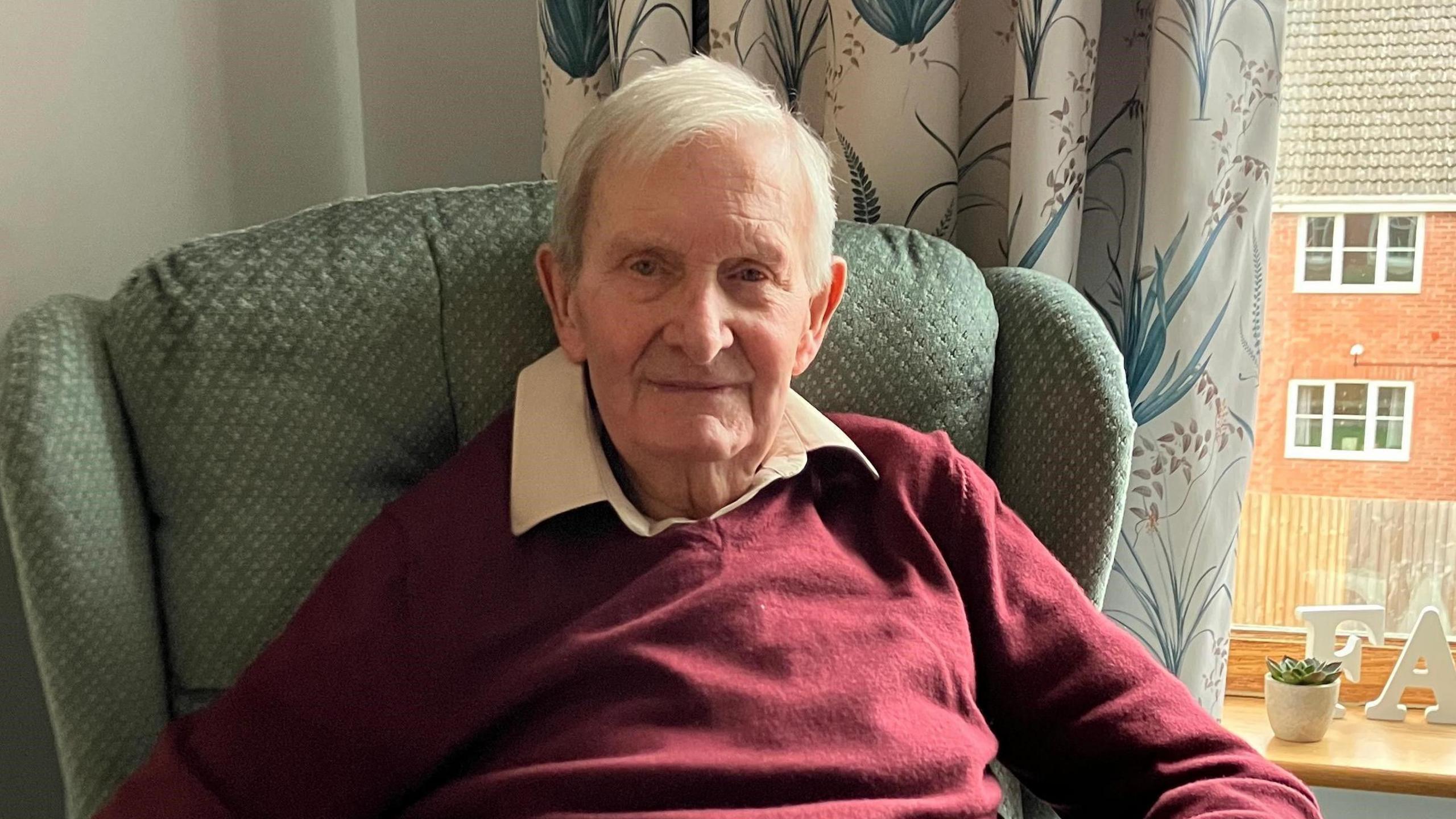 A man with a burgundy jumper and white hair looks into camera, sitting in a green armchair with a floral print curtain behind it. and a window through which another building can be seen.