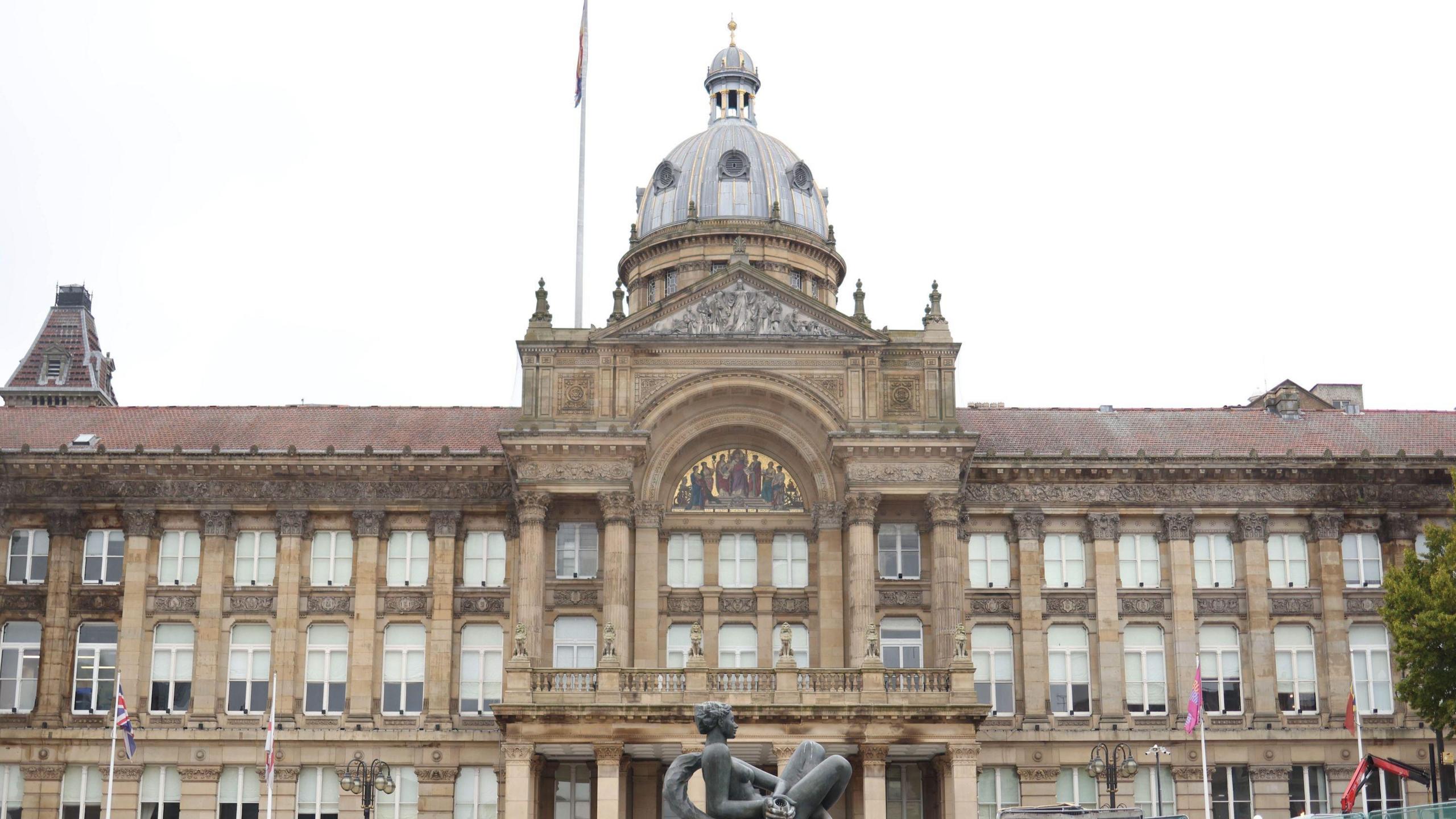 The exterior of the Birmingham City Council building, a Victorian-style stone building with a large central dome atop the pillared and arched entrance. There are wide steps and a fountain with a black sculpture of a person in front of it.