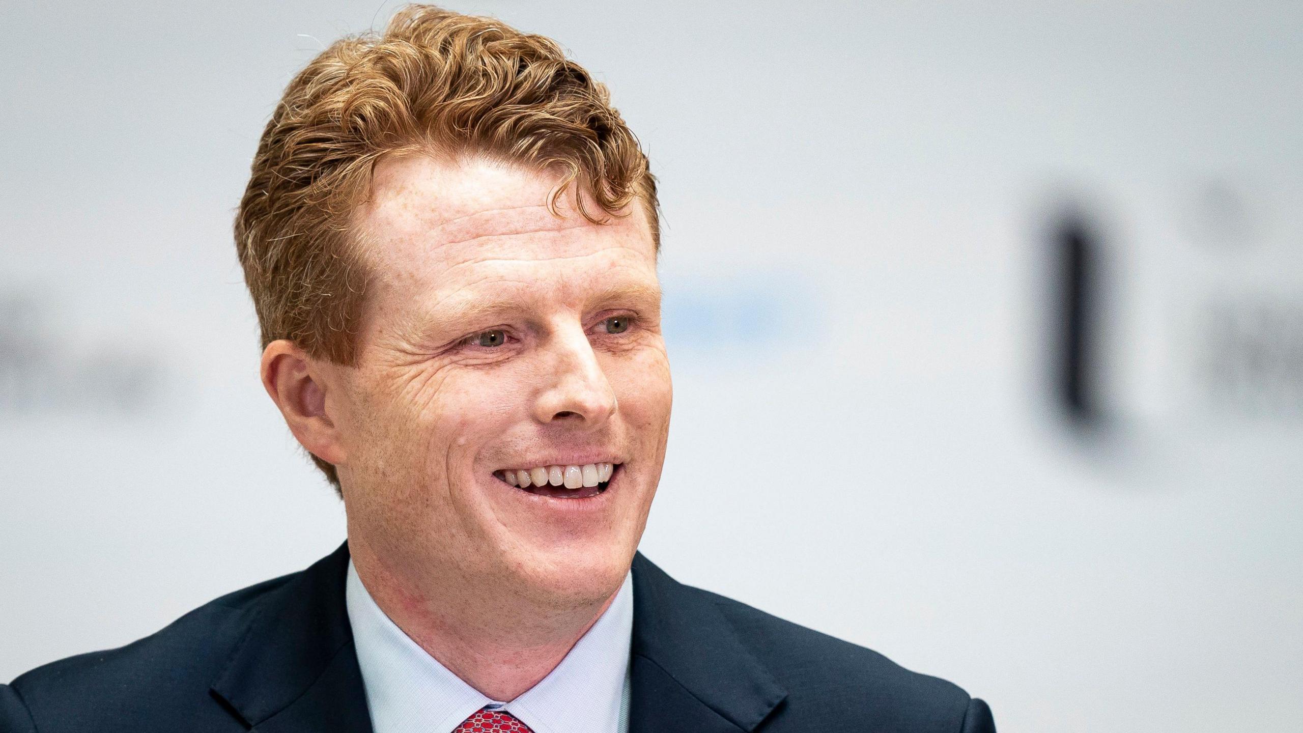 Joe Kennedy smiles. He has ginger hair and freckles. He is wearing a white shirt with a red tie and a blue blazer.  The background behind him is blurred.