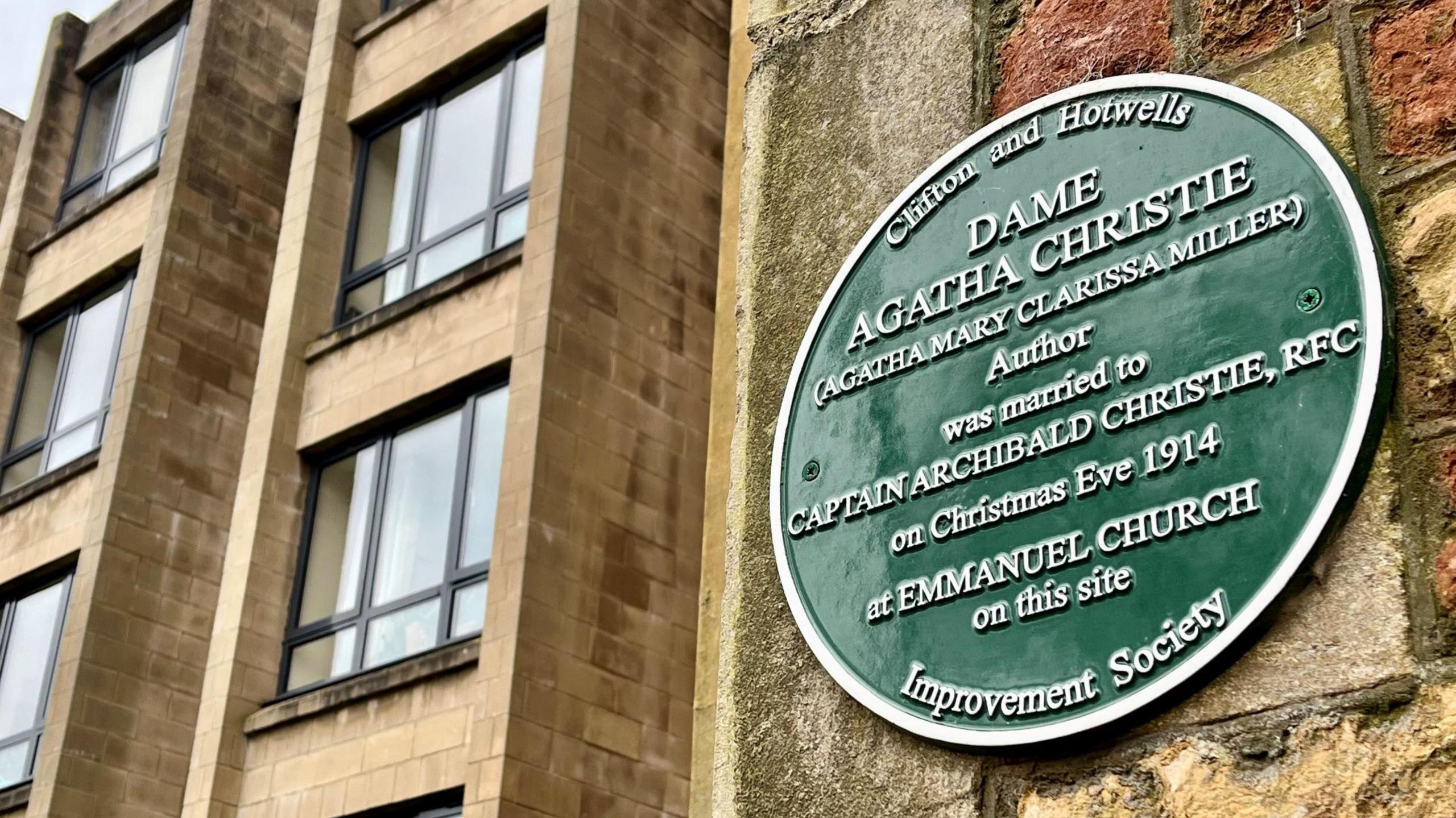 A large green plaque on the side of a stone wall in Bristol explains how the author Agatha Christie was married there in 2014. In the background is a more modern building with dark frames and stylish light brown bricks