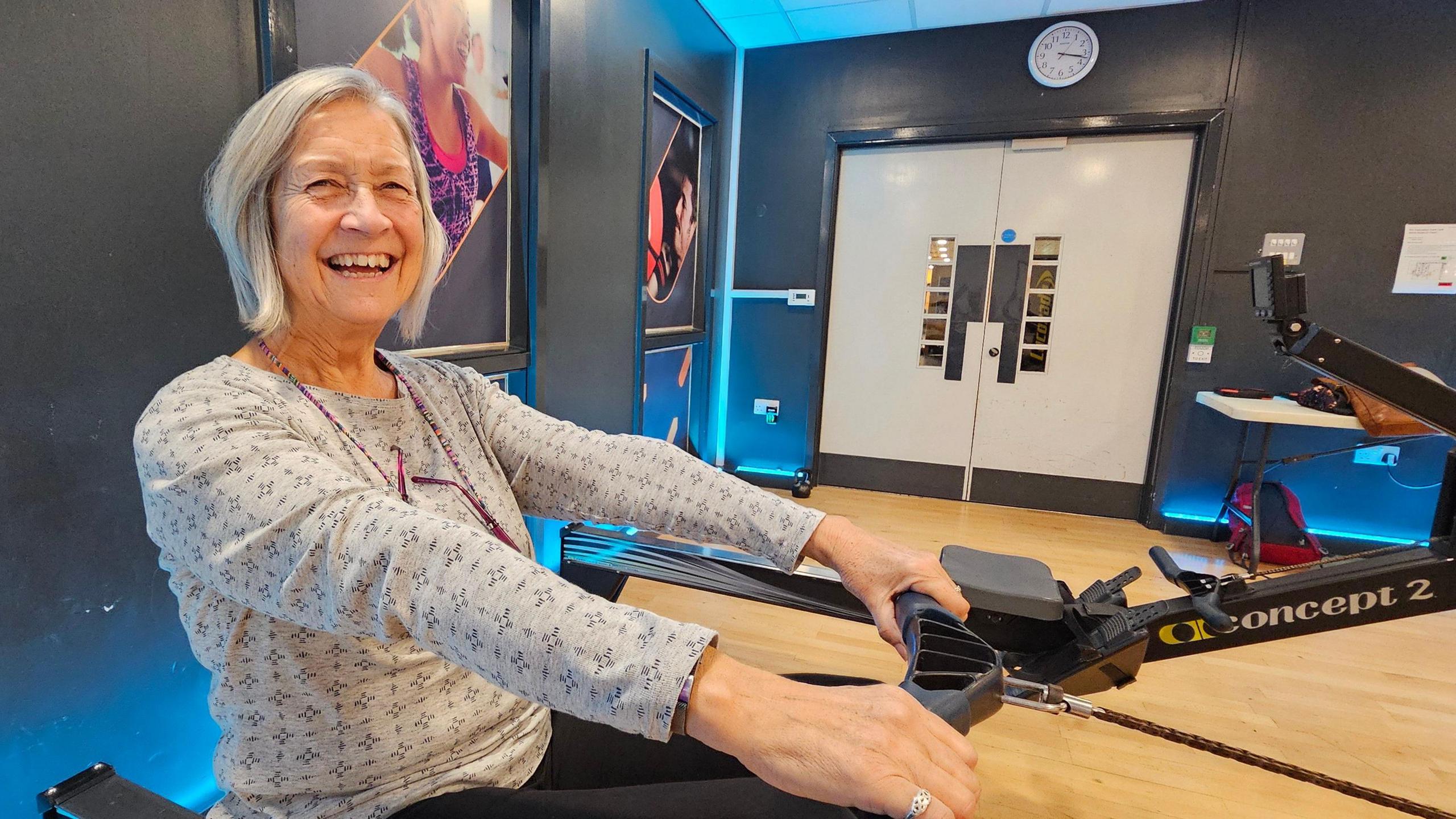 Pam Jackson sitting on a rowing machine. She has silvery bobbed hair to just above her shoulders. She's smiling broadly whilst wearing a grey, patterned top