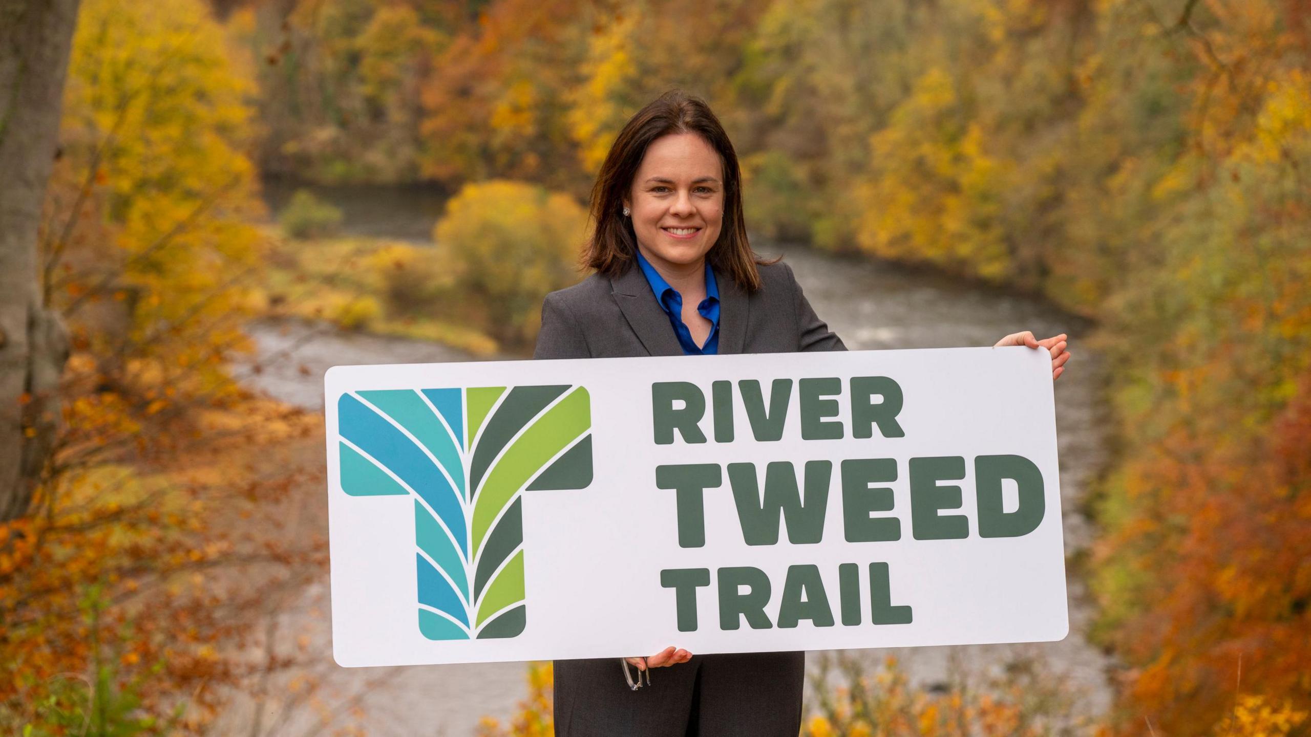 Deputy First Minister Kate Forbes holding up a new River Tweed Trail sign on a site overlooking a river and colourful autumnal trees