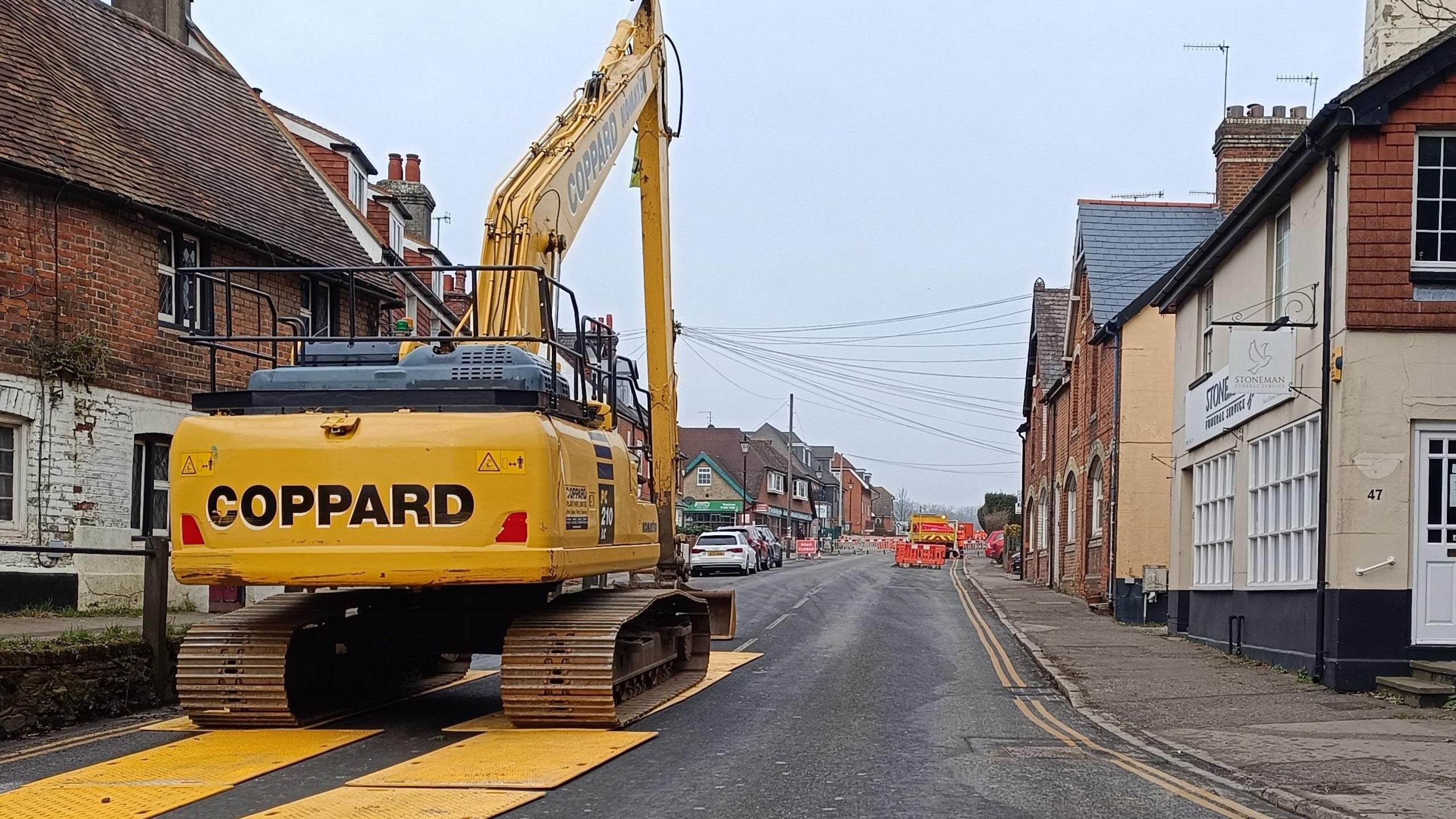 A yellow digger is parked at the top of a road with houses on either side. 