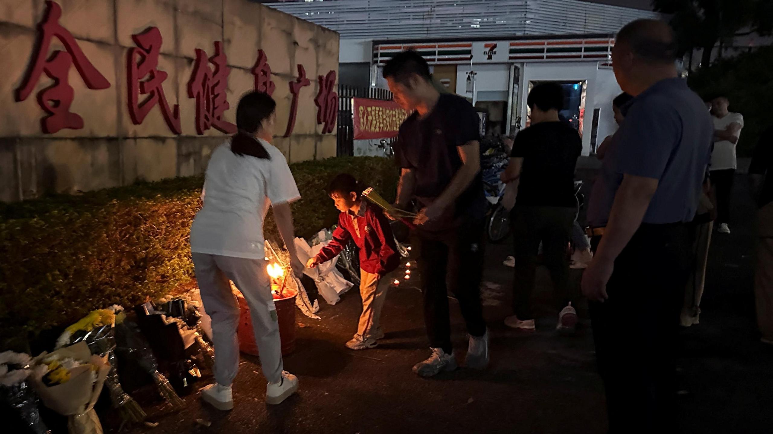 A small boy wearing a read top lays down flowers outside the stadium accompanied by two adults. More people watch from a bit further back, while the stadium and a 7-11 can be seen in the background