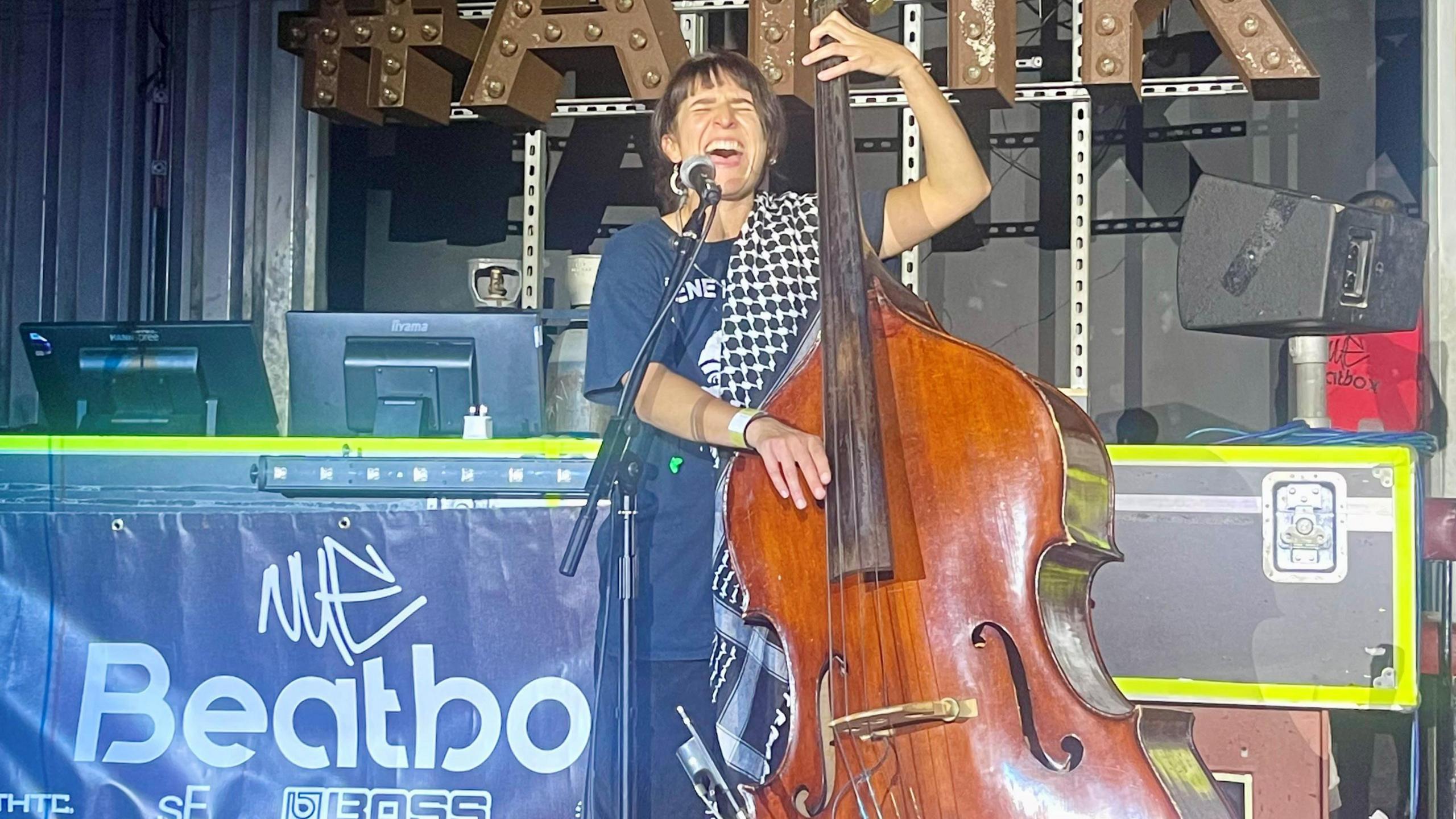 Former UK female beatboxing champion Bellatrix with her eyes closed performing at the microphone and playing a double bass. 