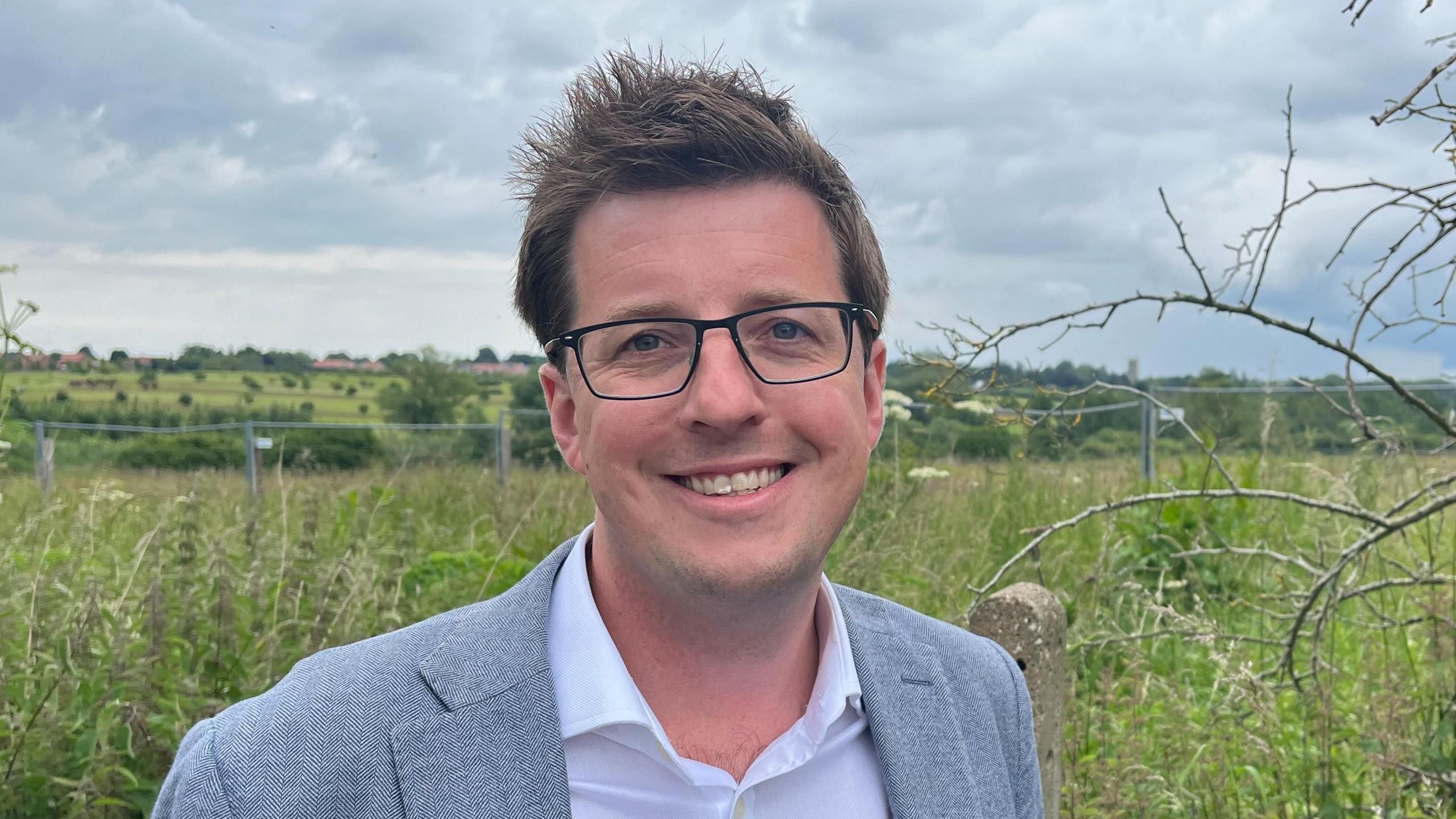 Iain Simpson wearing an open white shirt and suit jacket, a field behind him