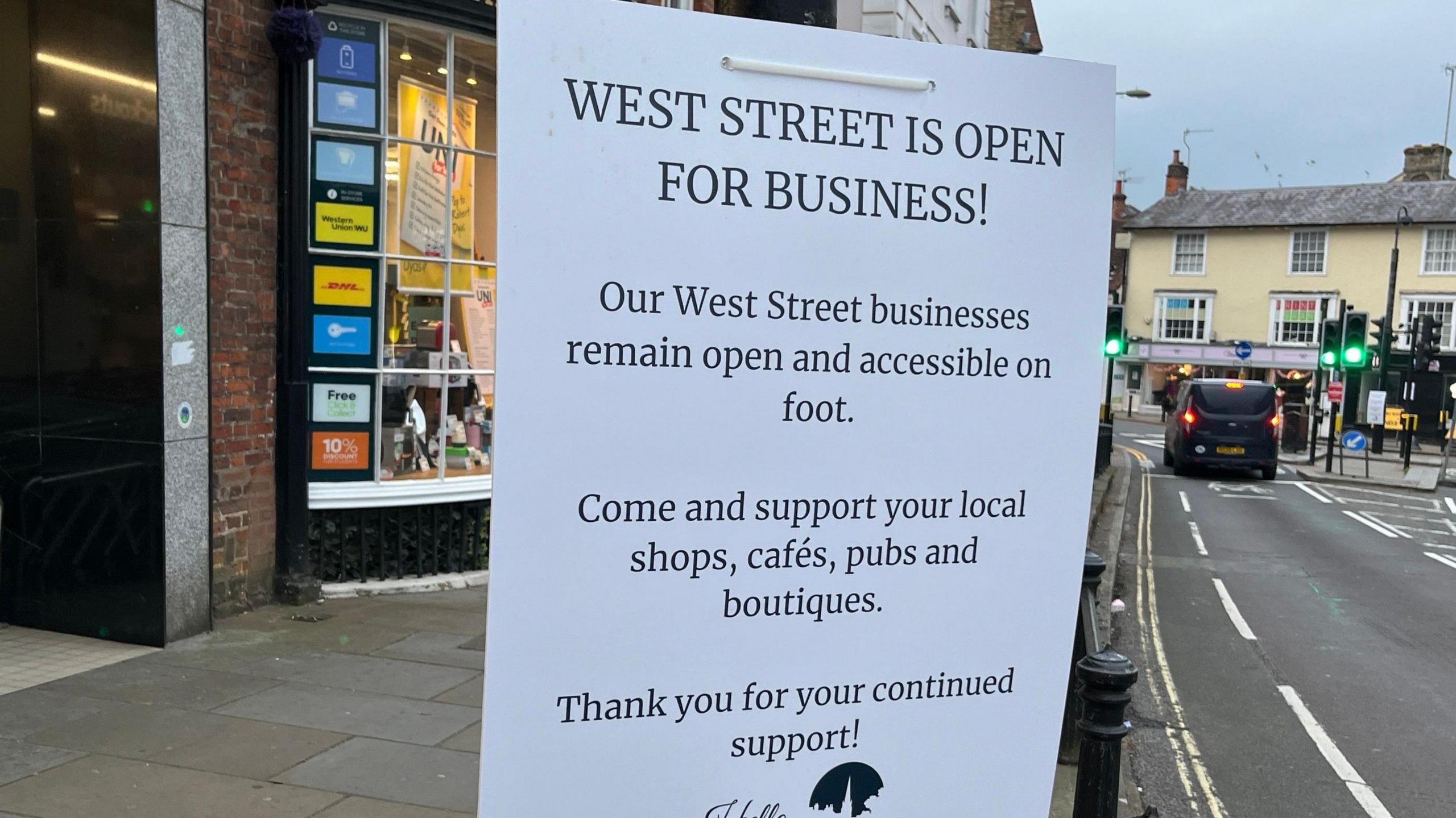 A laminated piece of paper tied to a post next to a road, which is on the right. The paper reads 'West Street is open for business! Our West Street businesses remain open and accessible on foot. Come and support you local shops, cafes, pubs and boutiques. Thank you for your continued support!'