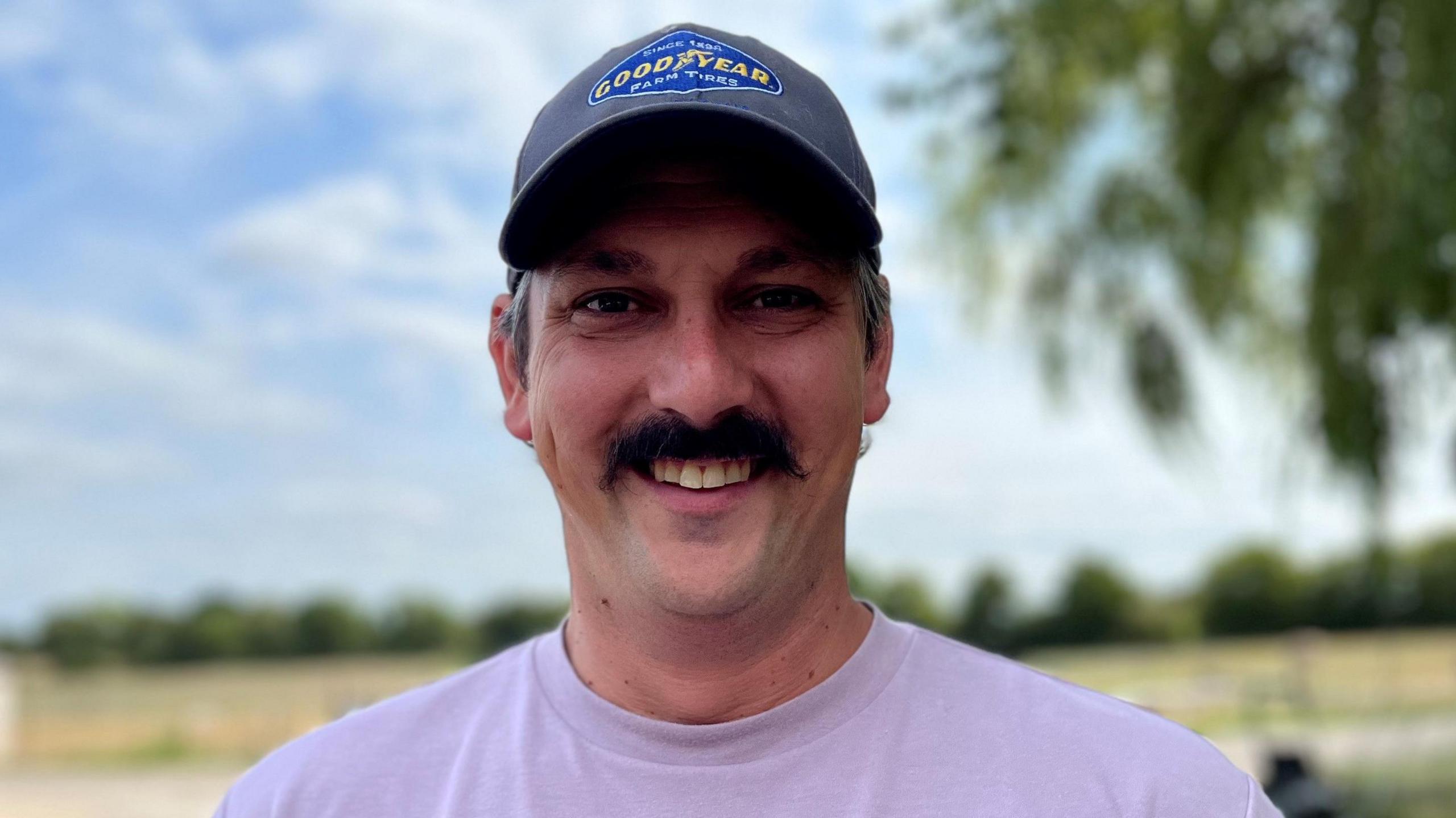 A farmer, who wears a cap and a lilac t-shirt, smiles outdoors.
