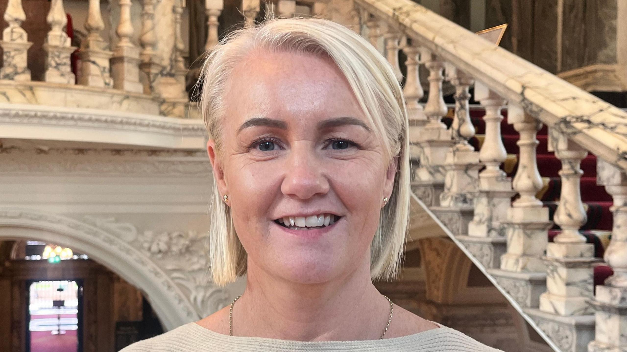 Tracy Kelly standing in Belfast City Hall, she is wearing a cream top and has blonde hair