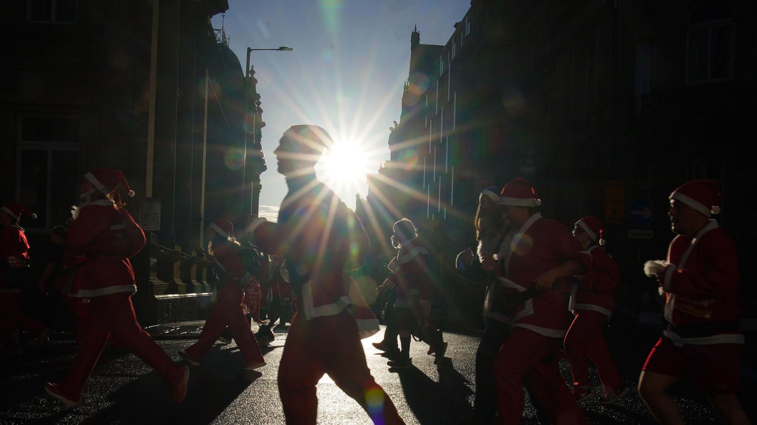 Glare of the rising sun shining through a street, creating a silhouette of people running past grand buildings in Santa suits.