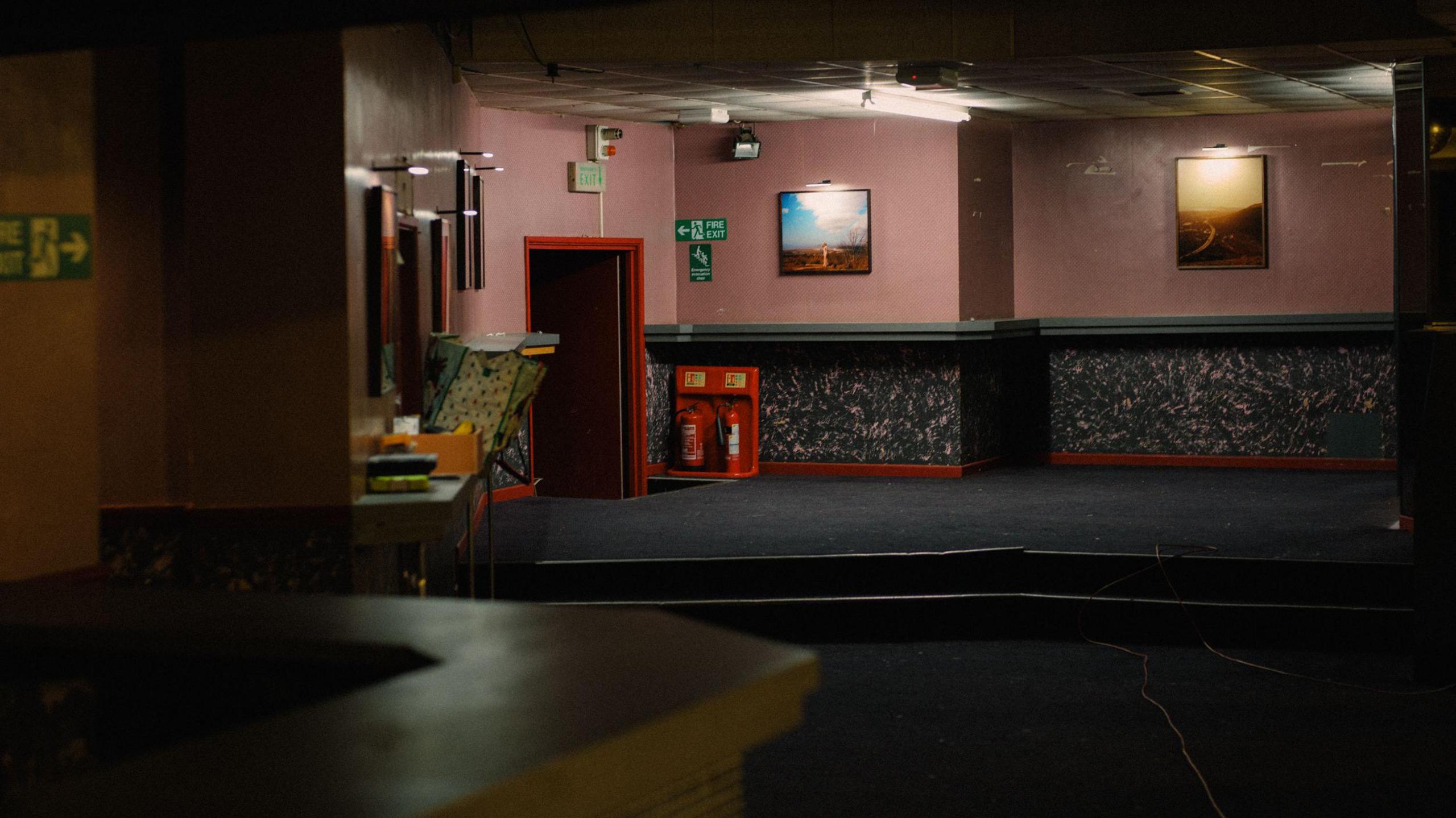 A dark basement room with pictures hanging on the far wall. To the left of the wall is a fire exit sign and beneath those, fire extinguishers. In the foreground and to the left is a bar