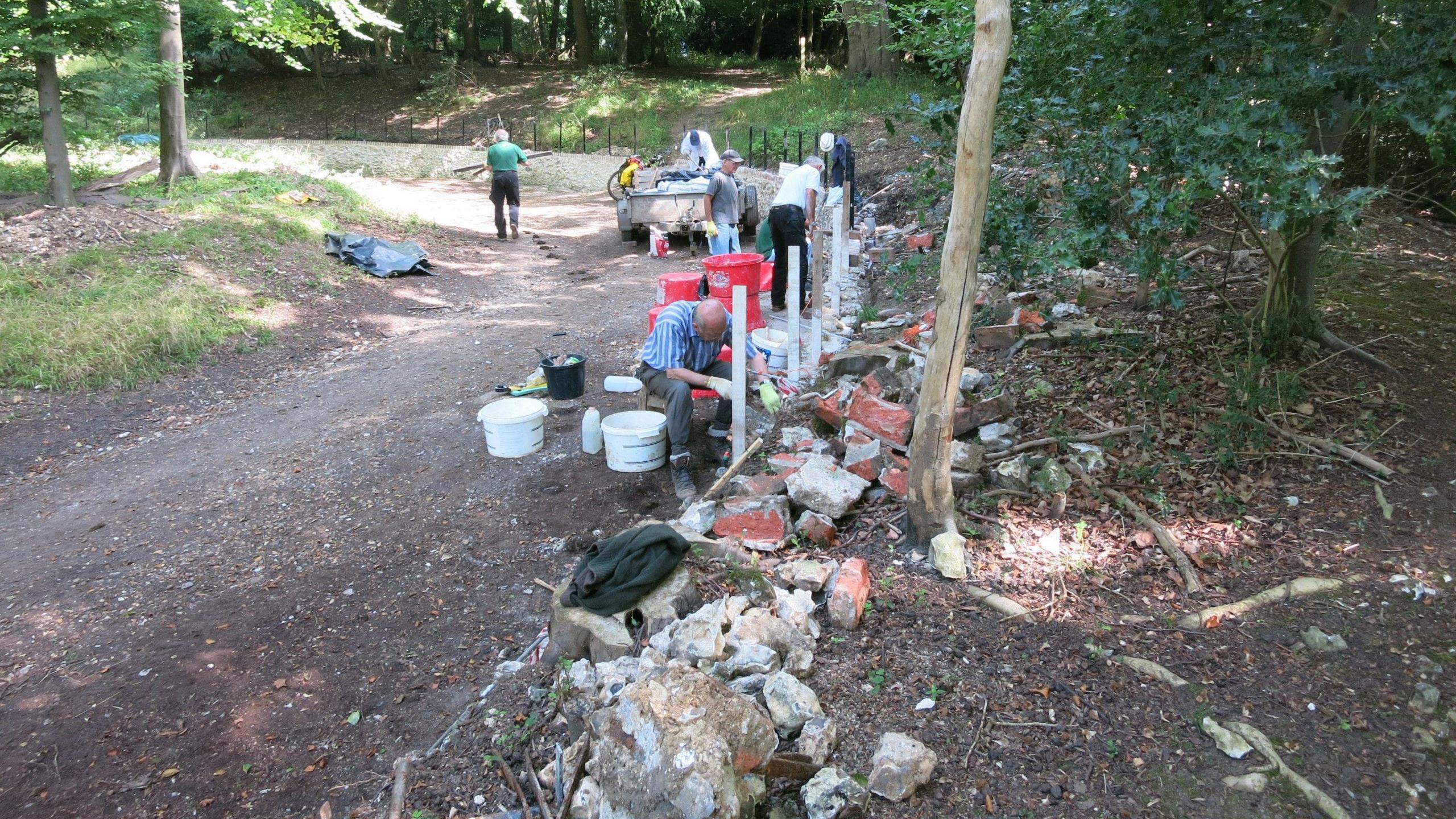 Four volunteers rebuilding a brick wall, in a forest area with trees, shrubs and a path. 