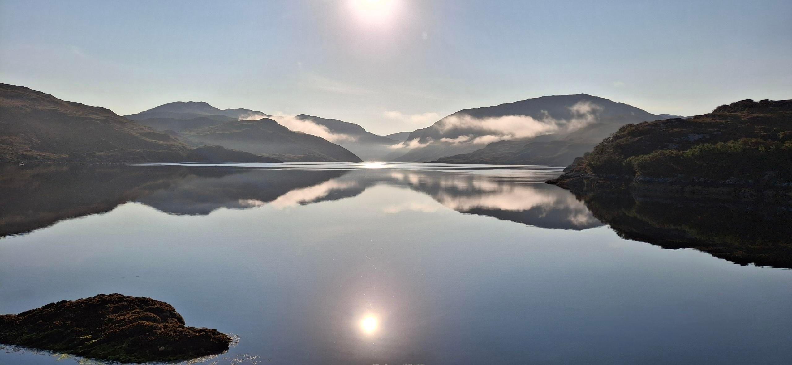 A mountain range, which is reflected on a loch, with the sun high in the picture