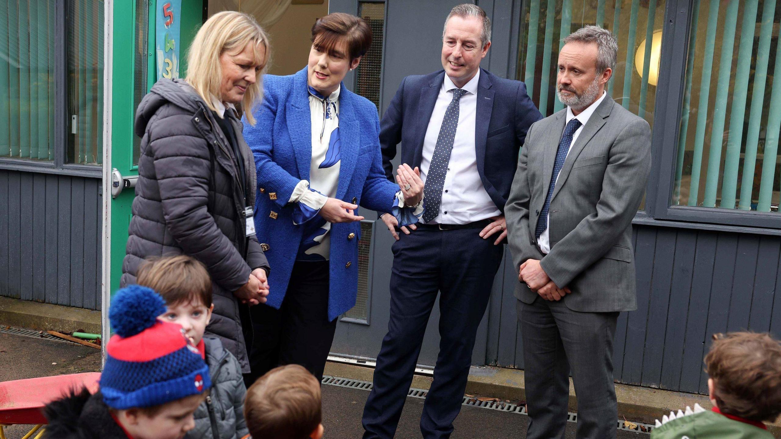 Paul Givan, wearing a dark blue suit, white shirt and dark tie stands with his hands on his hips outside a school.  Standing beside him is Irish Education Minister Norma Foley wearing a bright blue jacket and white and blue blouse.  She is talking and gesturing with her hands.  They are talking to a woman with blonde hair and a grey-haired man in a suit.   Four children are in the foreground, partially out of view.