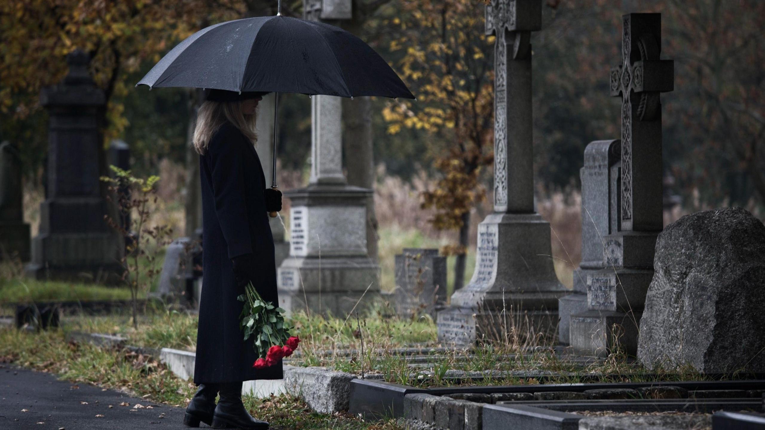Stock photo of a lady bringing roses to a grave in a cemetery. She is wearing all black, has long blonde hair and is holding a large black umbrella above her head.